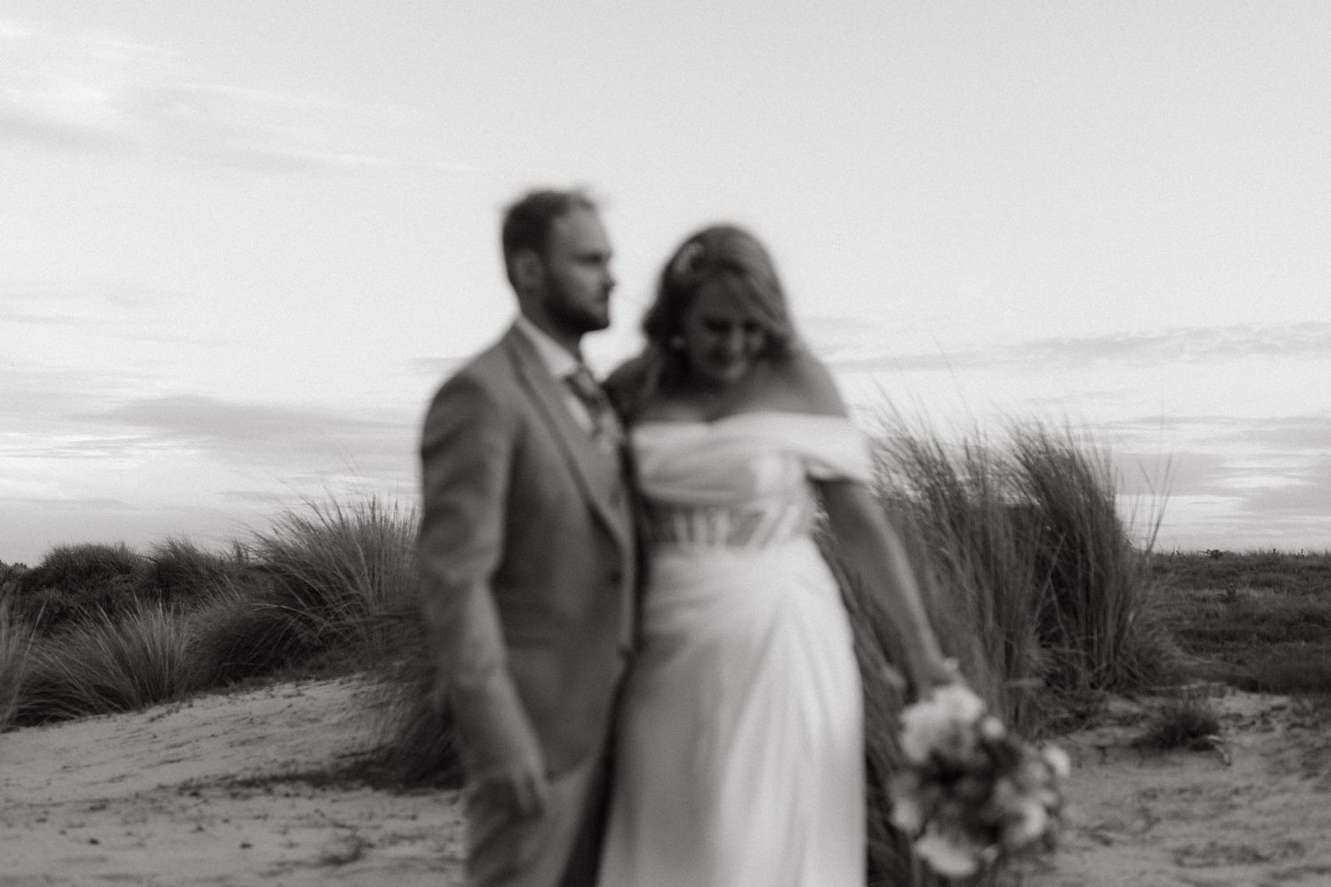 A romantic black and white wedding photo of a couple standing together against beach grass at sunset.
