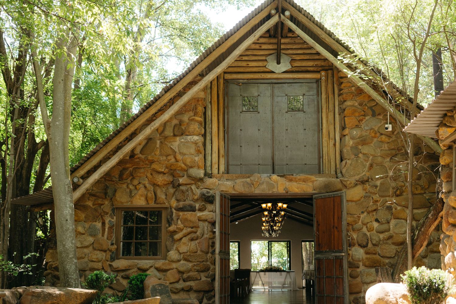 A rustic stone chapel with wooden beams and gray doors sits nestled among green trees in a forest setting.