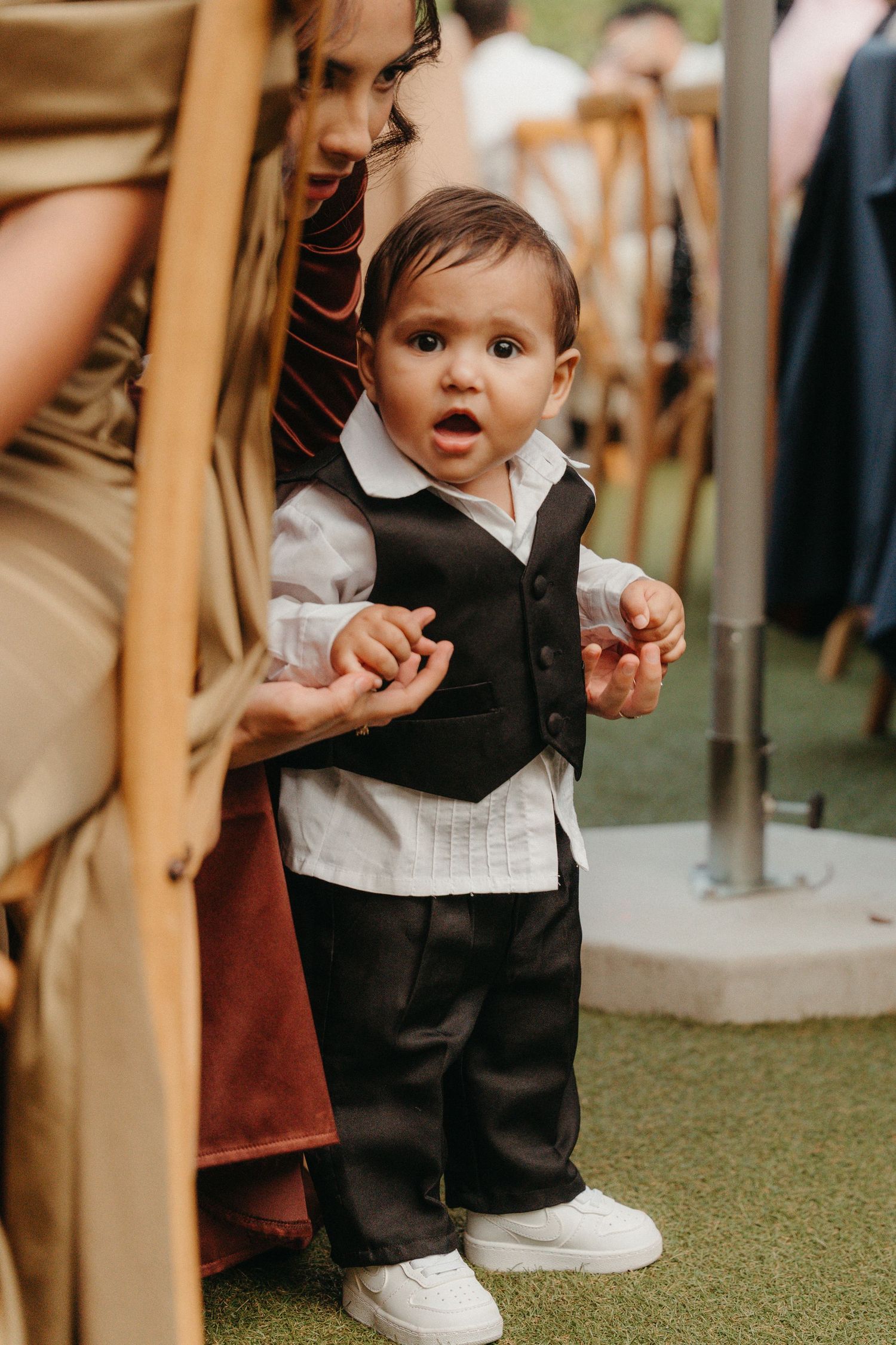 Adorable baby boy in a formal black vest and white shirt stands at a wedding reception on green grass.