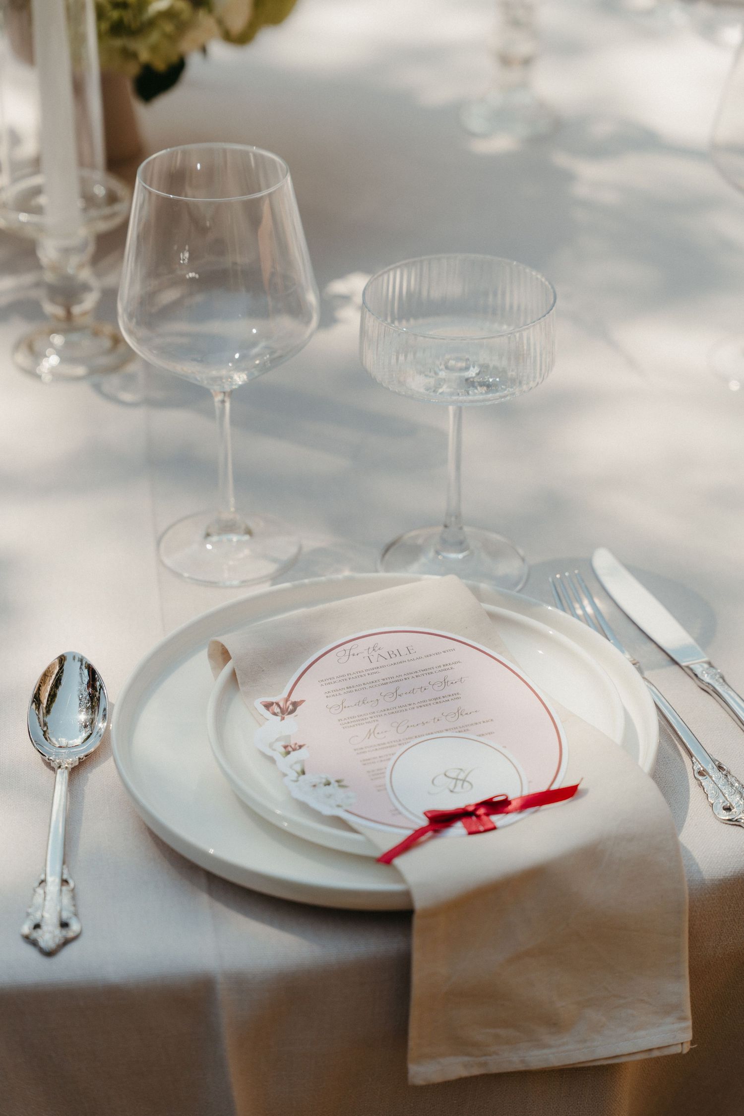 Elegant wedding table setting with white plates, circular menu card, red ribbon, silver cutlery, and crystal glassware on white linen.