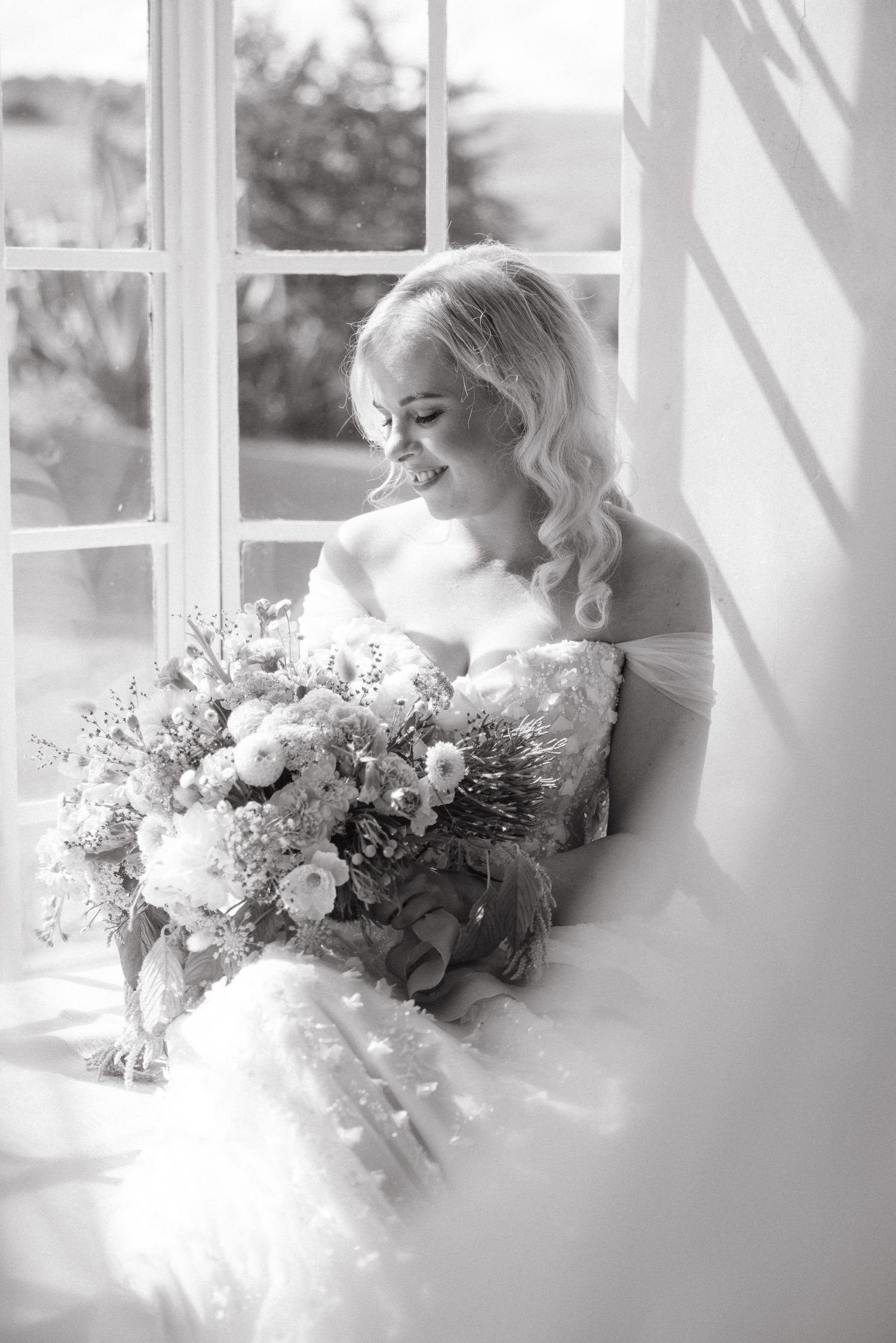 Black and white photo of a smiling bride in a strapless gown holding a floral bouquet while seated by a sunlit window.