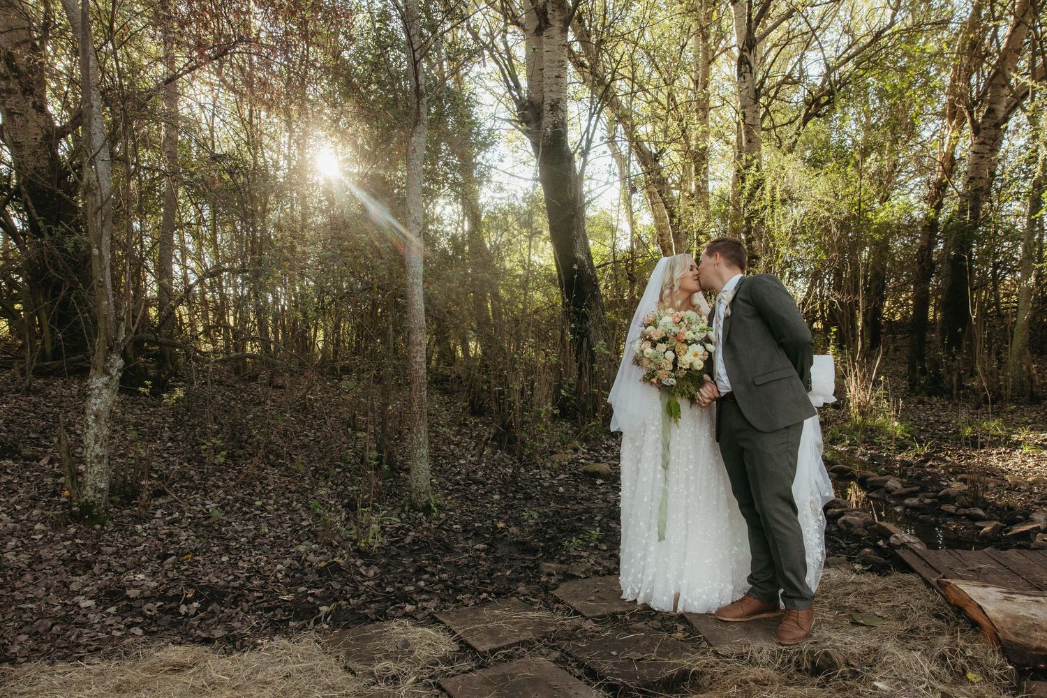 Bride and groom sharing a kiss in a sunlit forest, bride holding a bouquet in a white gown and veil.