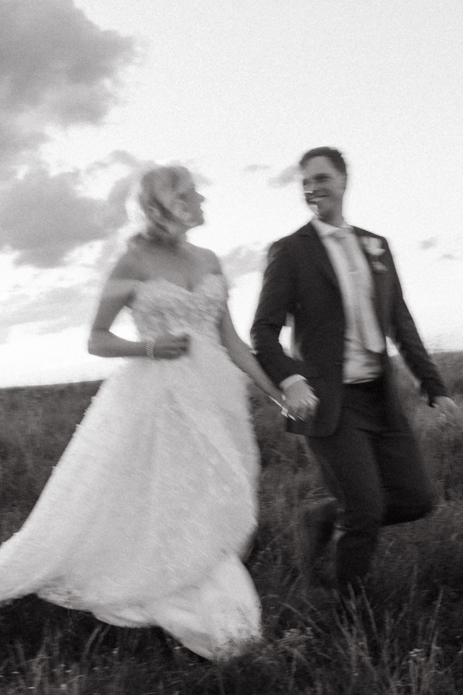 Black and white photo of a joyful bride and groom holding hands and running through a field on their wedding day.