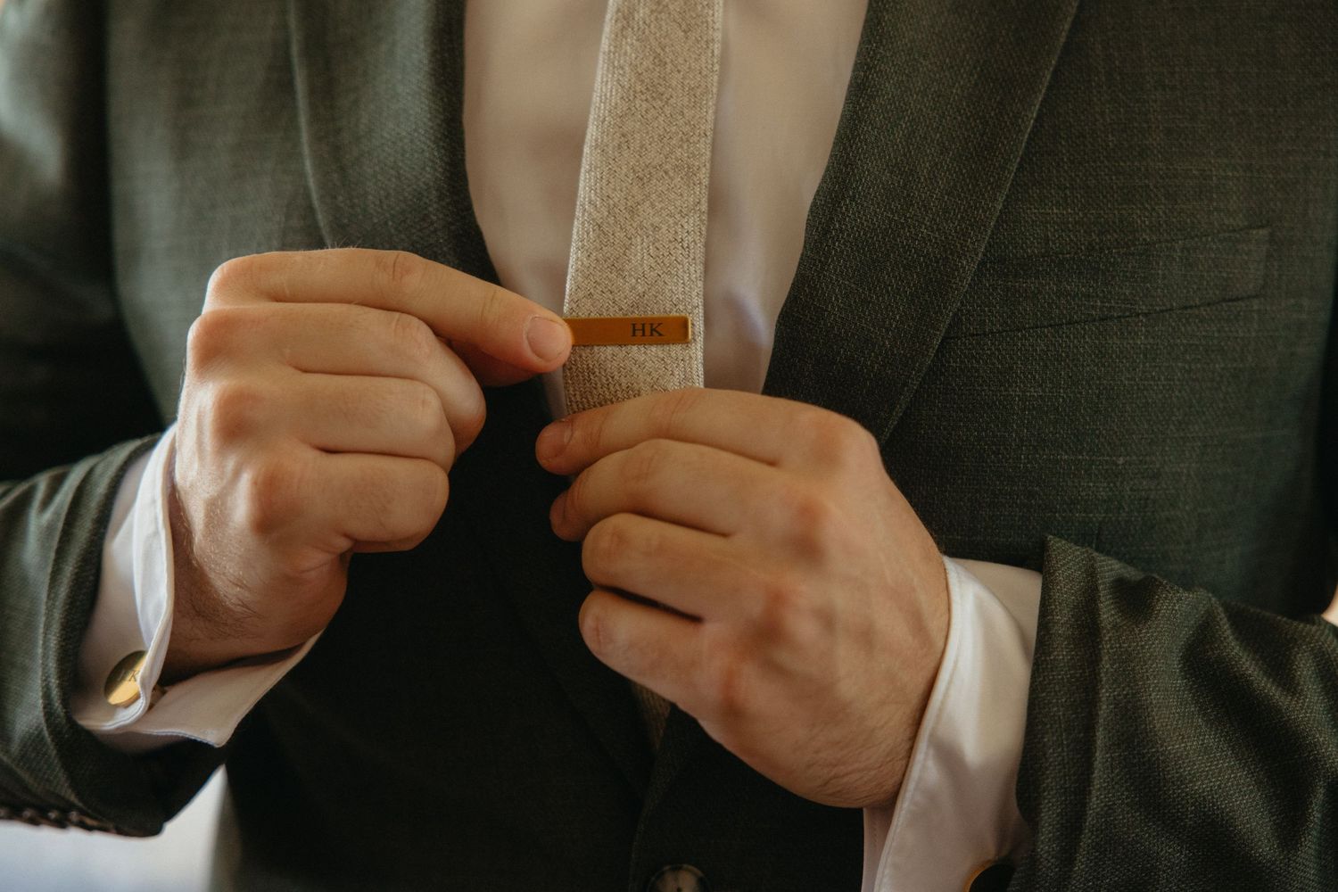 Man in gray suit adjusting a gold tie clip on a beige tie, close-up shot of hands and chest.