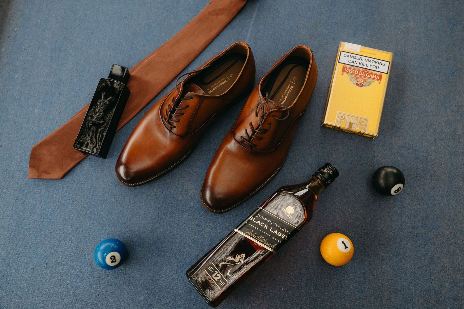 Brown leather oxford shoes with a tie, Johnnie Walker Black Label whiskey, cigars, and billiard balls on blue carpet.