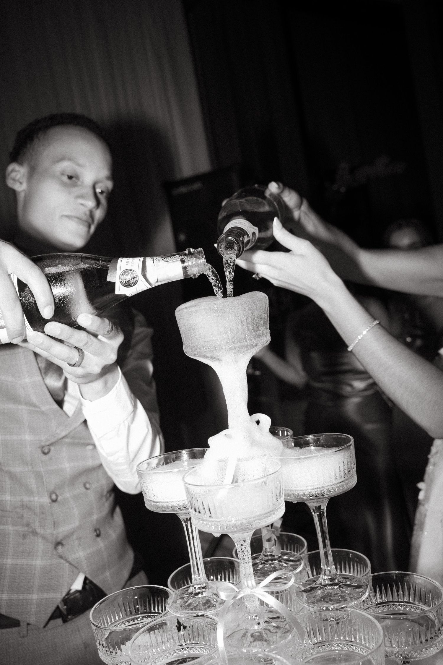Black and white photo of a bride and groom pouring champagne into a tiered glass tower at their wedding reception.