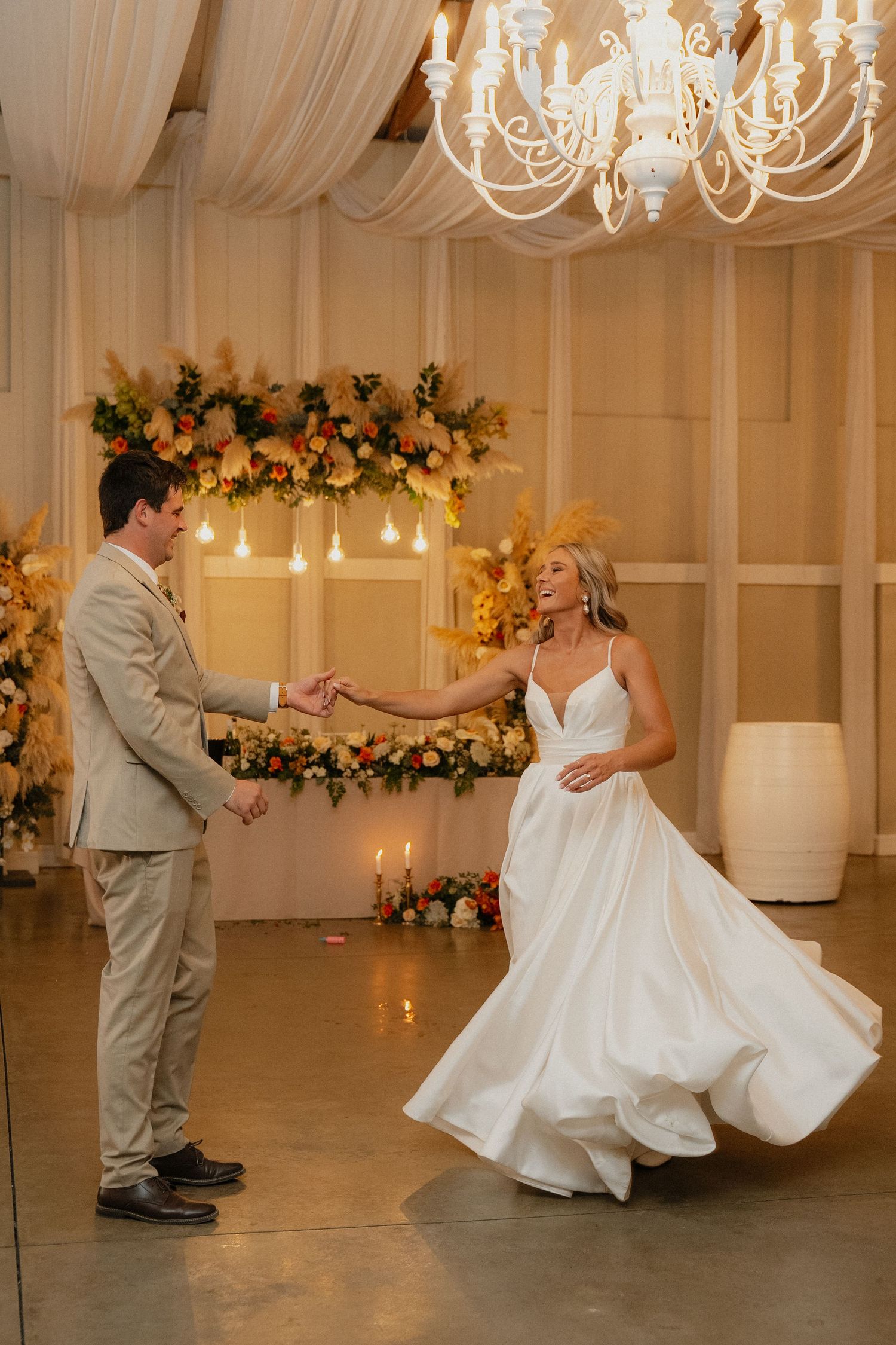 Bride and groom share their first dance under a chandelier, surrounded by warm floral arrangements and candlelight.