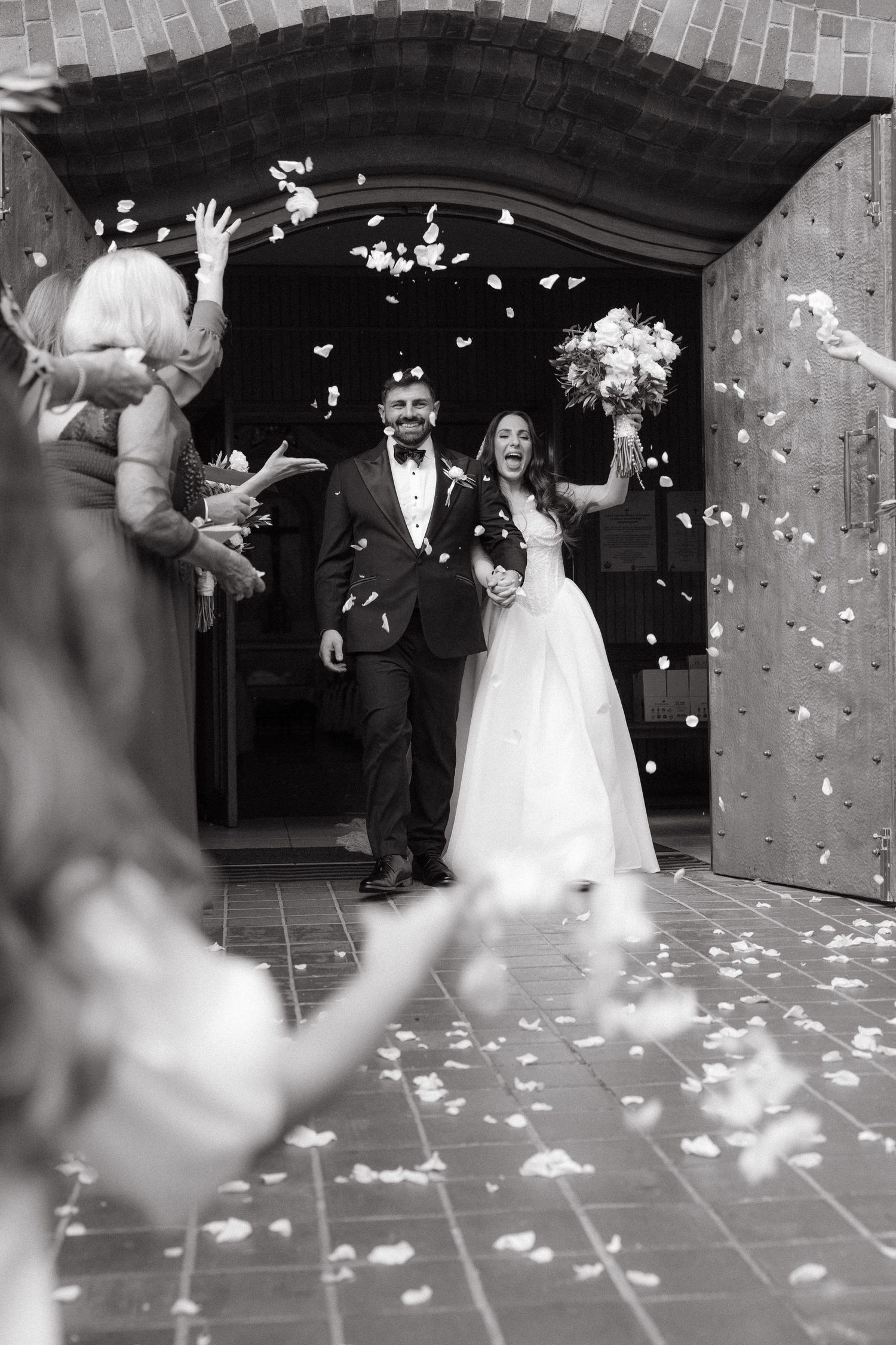 Black and white photo of a joyful bride and groom exiting Rosebank Catholic Church as guests toss flower petals in celebration.