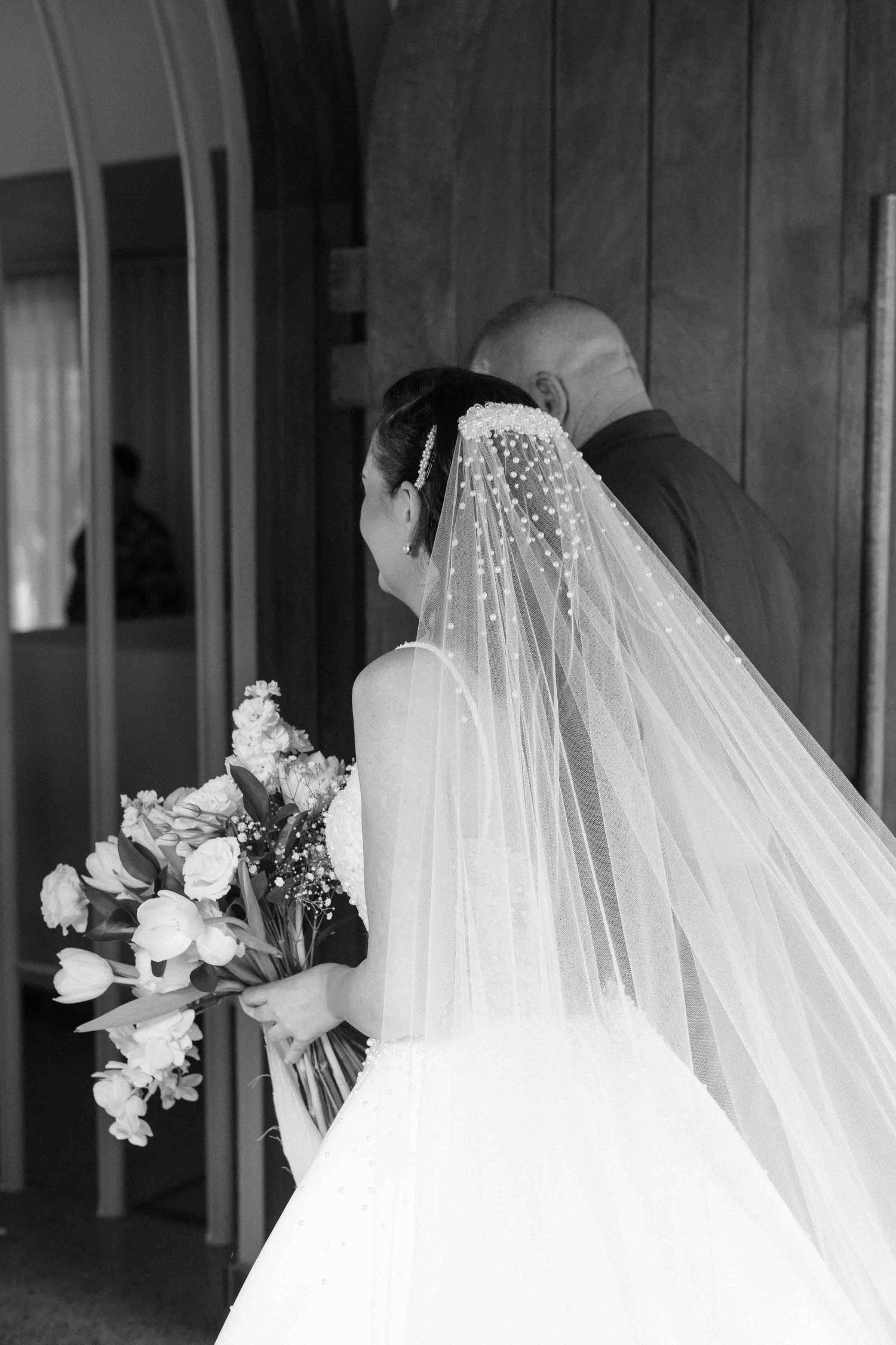 Black and white photo of a bride in a veil holding a bouquet, standing with a bald man at a church door.