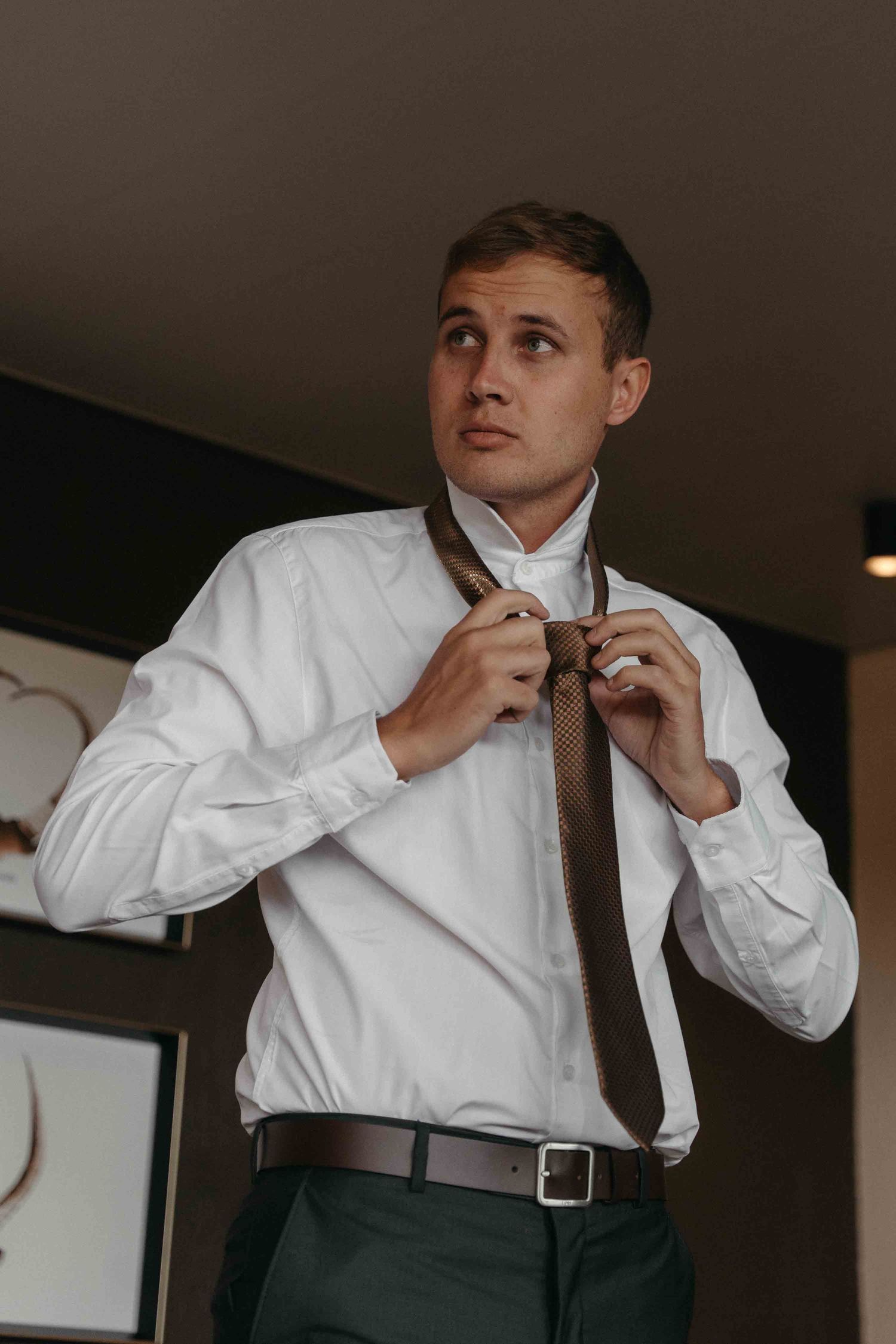Young man in white dress shirt adjusting brown knit tie, wearing green trousers and brown belt indoors.
