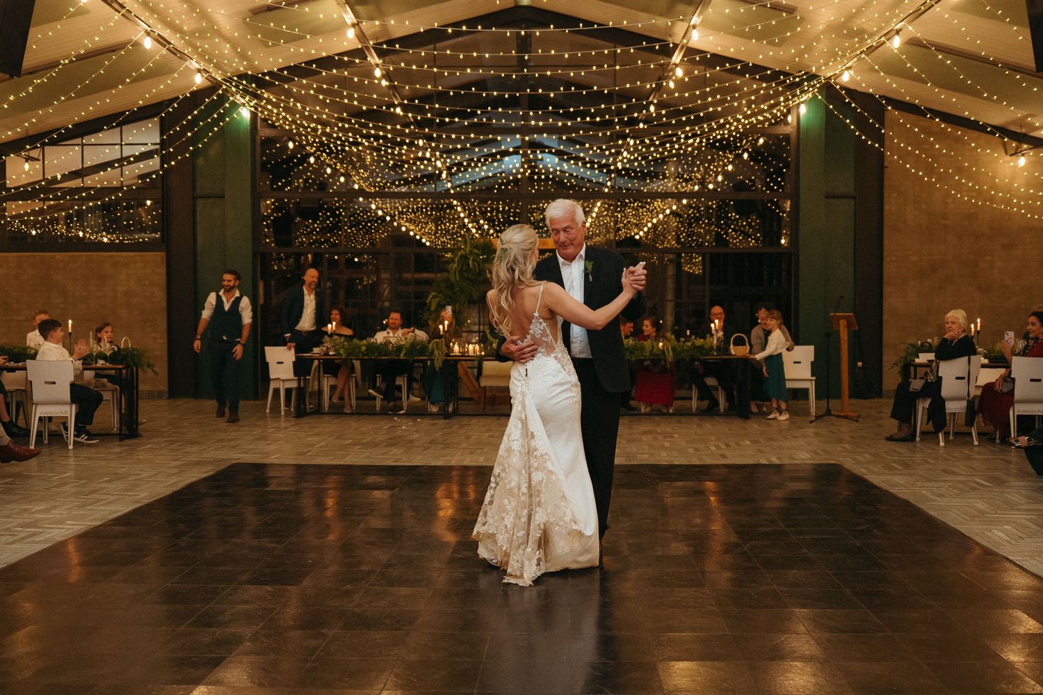 Bride and groom share first dance under twinkling fairy lights at elegant indoor wedding reception venue.