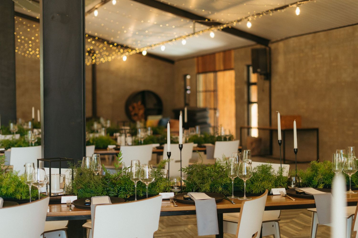 Elegant event venue with long wooden tables, white chairs, candles, greenery centerpieces, and string lights overhead.