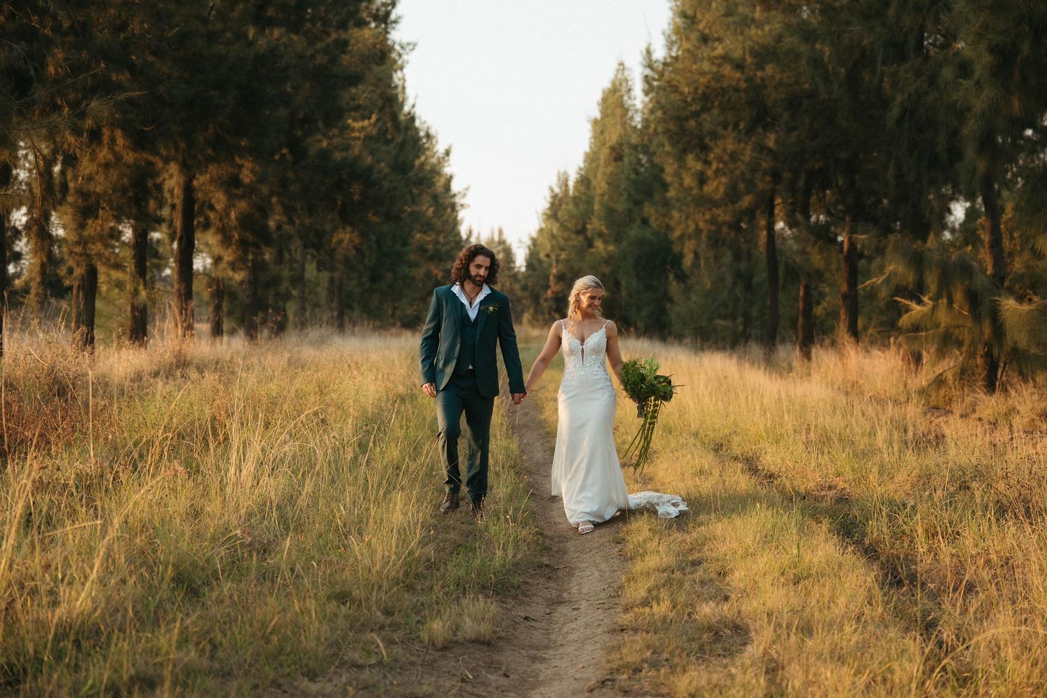 Bride and groom walking hand in hand down a rustic dirt path surrounded by tall trees and golden grass.