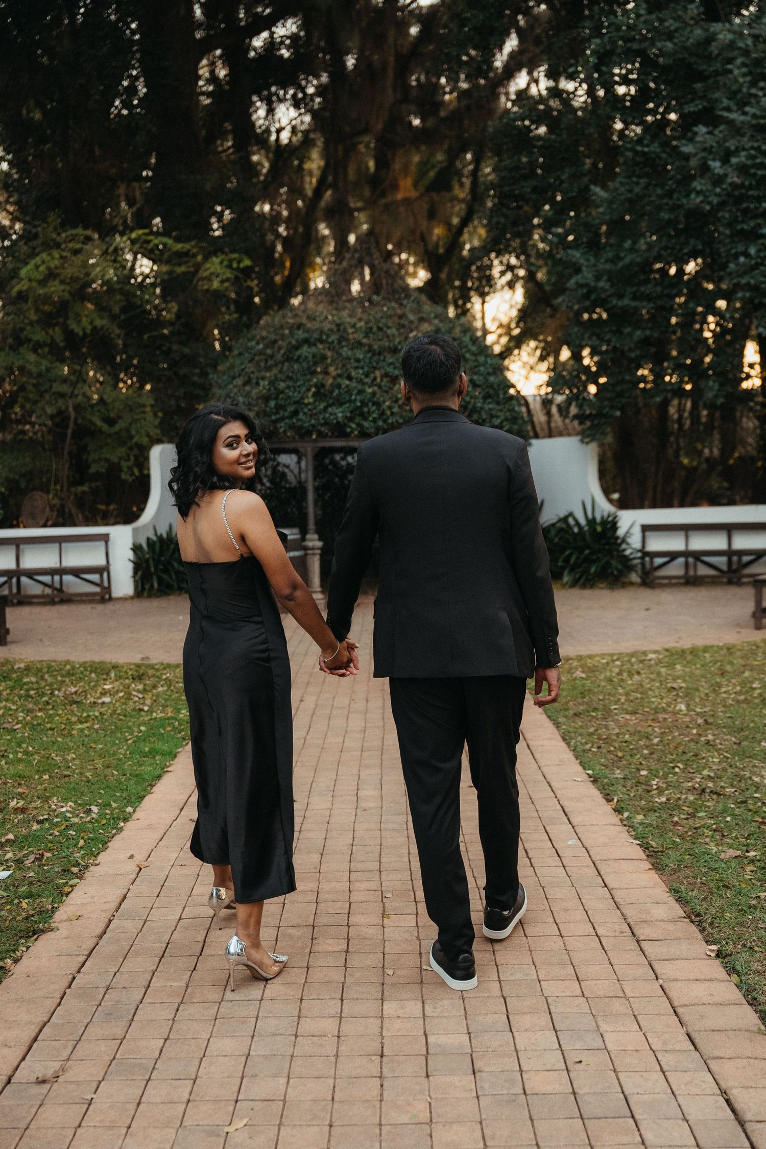 Couple in black formal attire holding hands walking on a brick path in a lush garden at sunset.