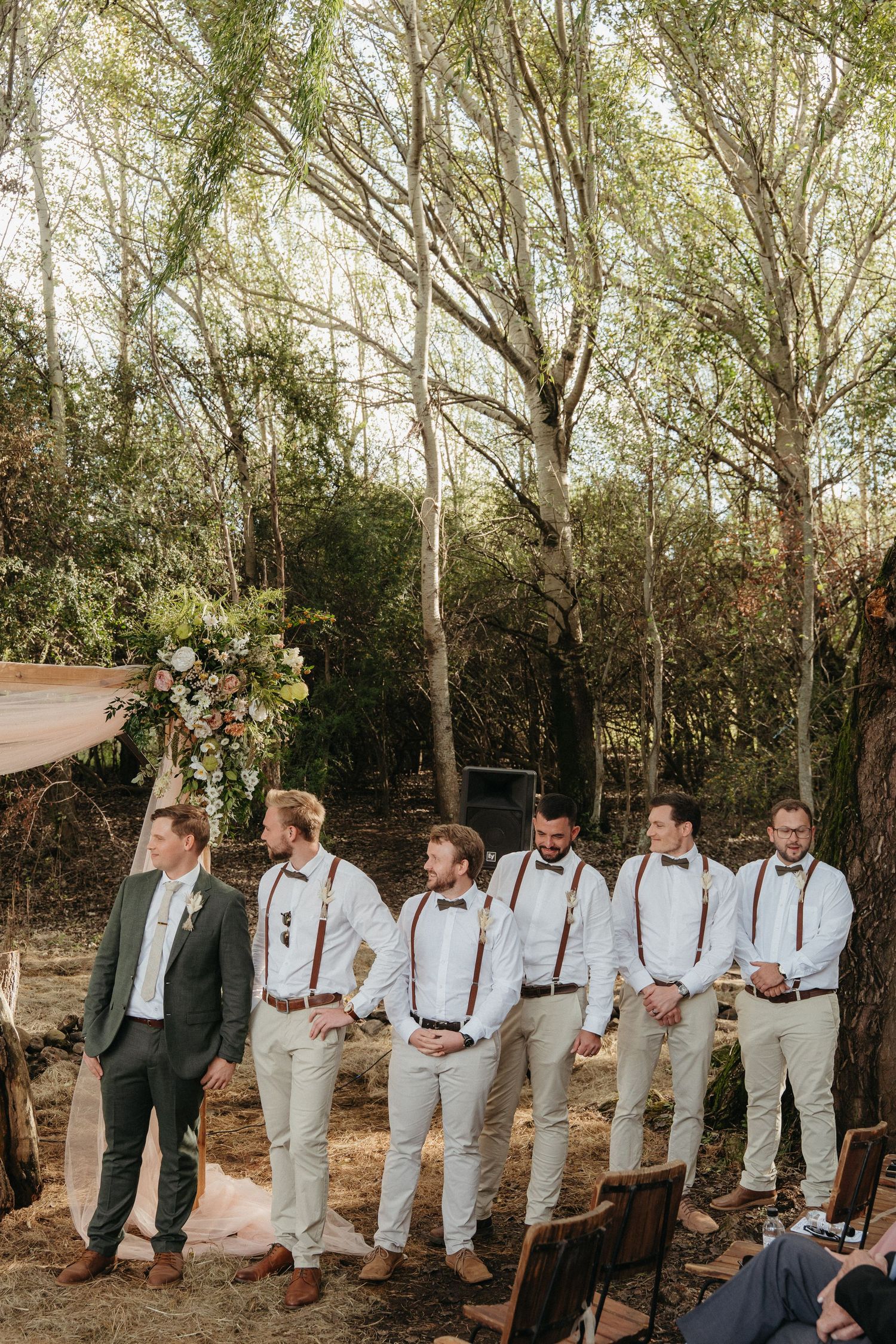 Groom with groomsmen in suspenders at an outdoor rustic woodland wedding ceremony.