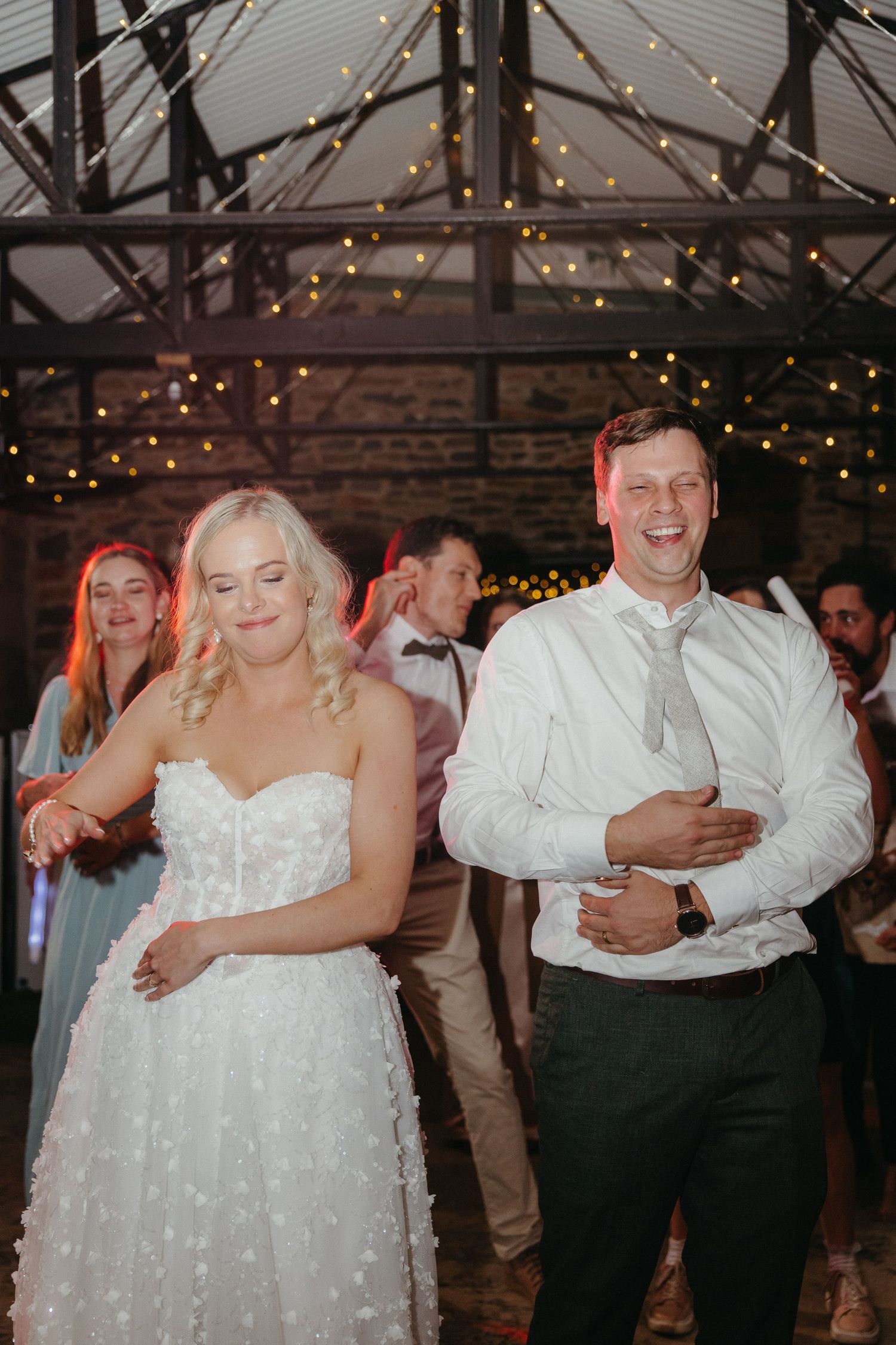 Bride and groom laughing and dancing together at their wedding reception under warm fairy lights.