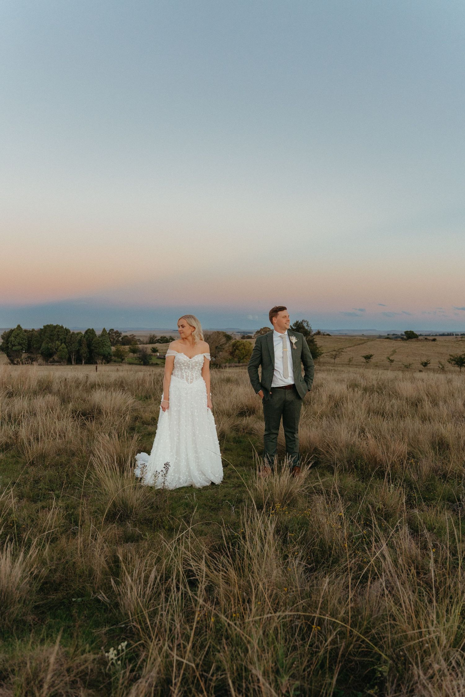 Bride in white lace gown and groom in dark suit stand in golden grass field at sunset.