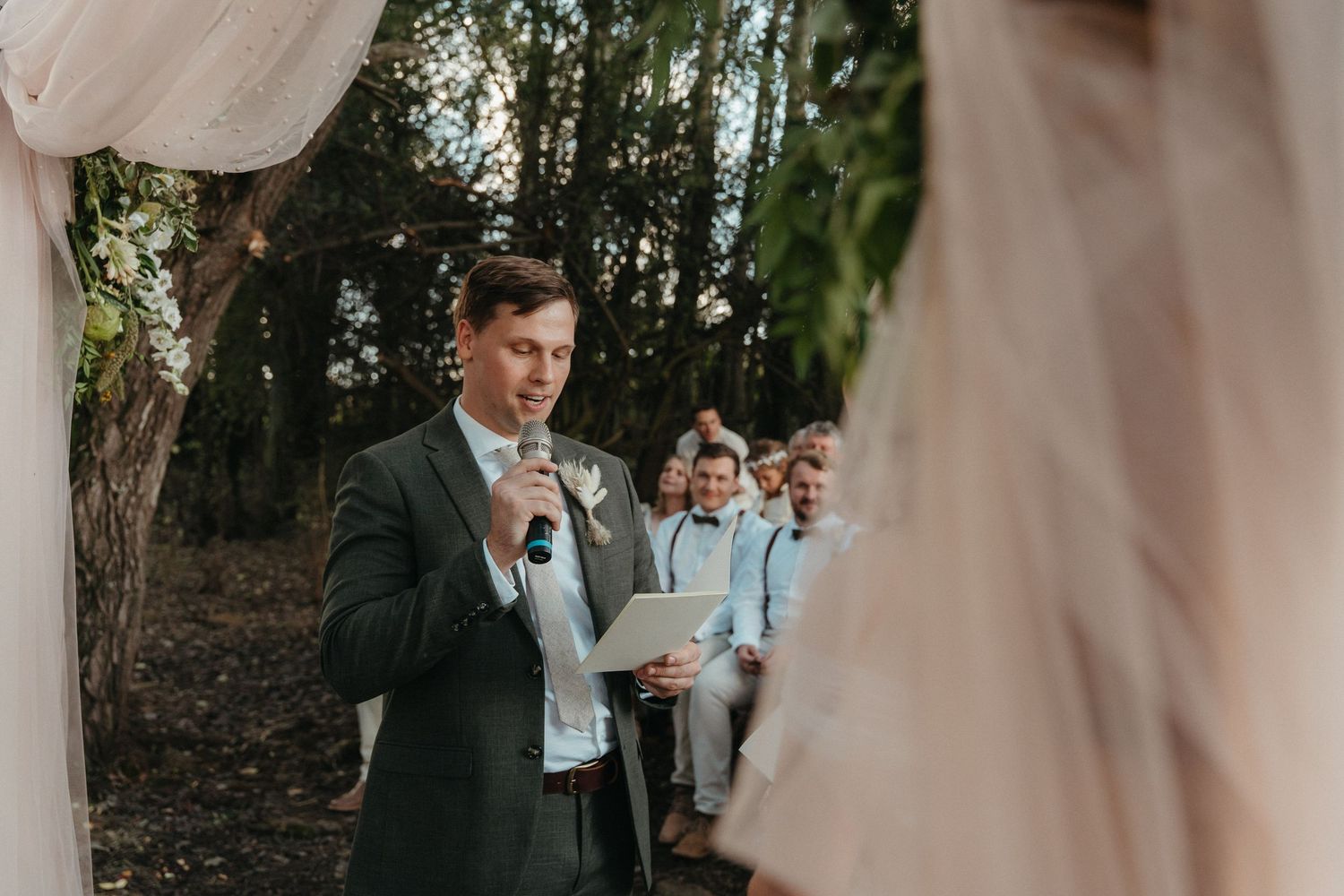 Groom reads vows at outdoor woodland wedding ceremony with groomsmen standing in background.