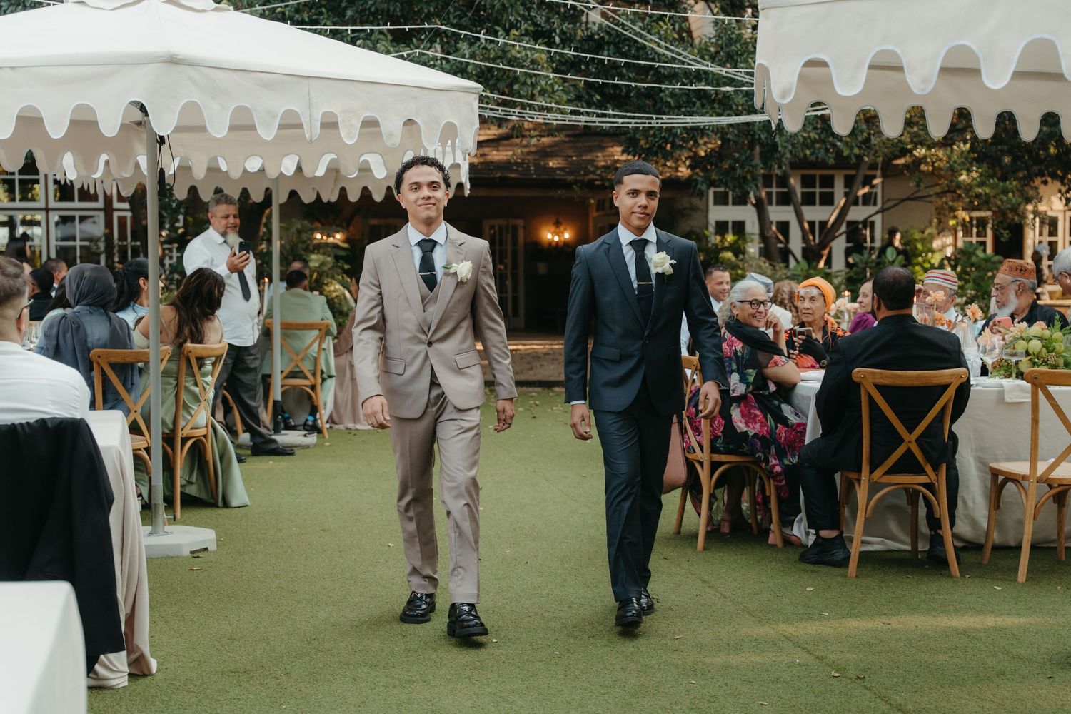 Two grooms walk down the aisle at an elegant outdoor wedding reception with white tents and seated guests.