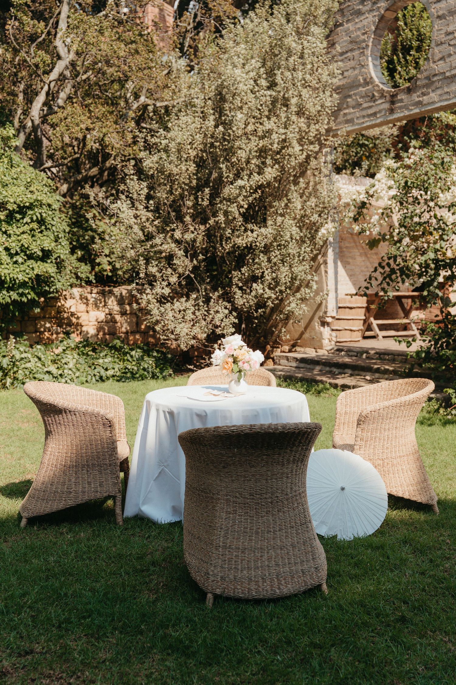 Wicker chairs surround a white-clothed table with flowers in a lush garden setting.