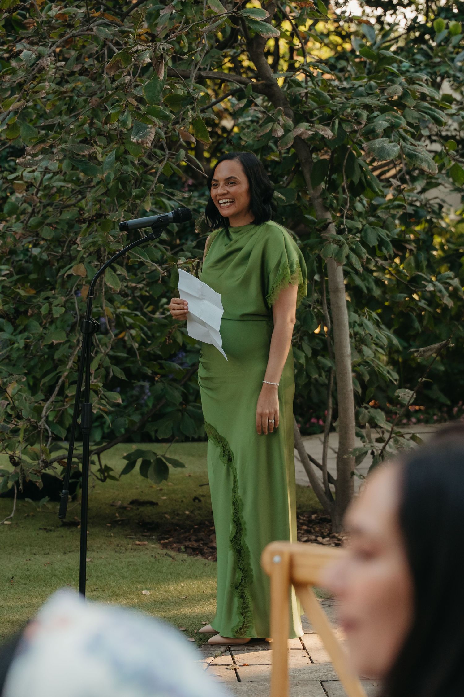 Woman in green jumpsuit smiling while giving a speech outdoors at a garden event, holding papers near a microphone.