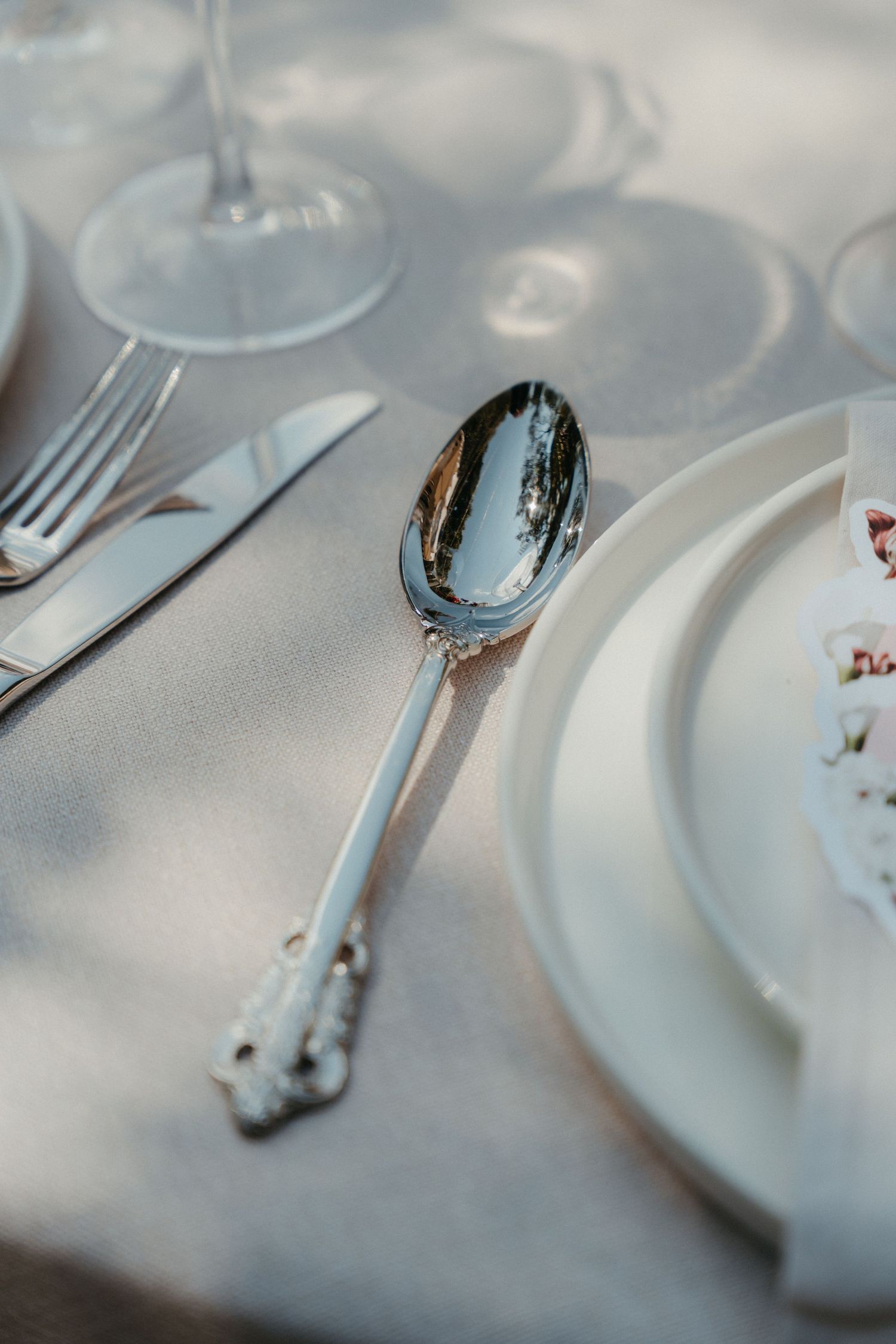 Elegant silver spoon and cutlery set on a white linen tablecloth with crystal glasses and white plates.