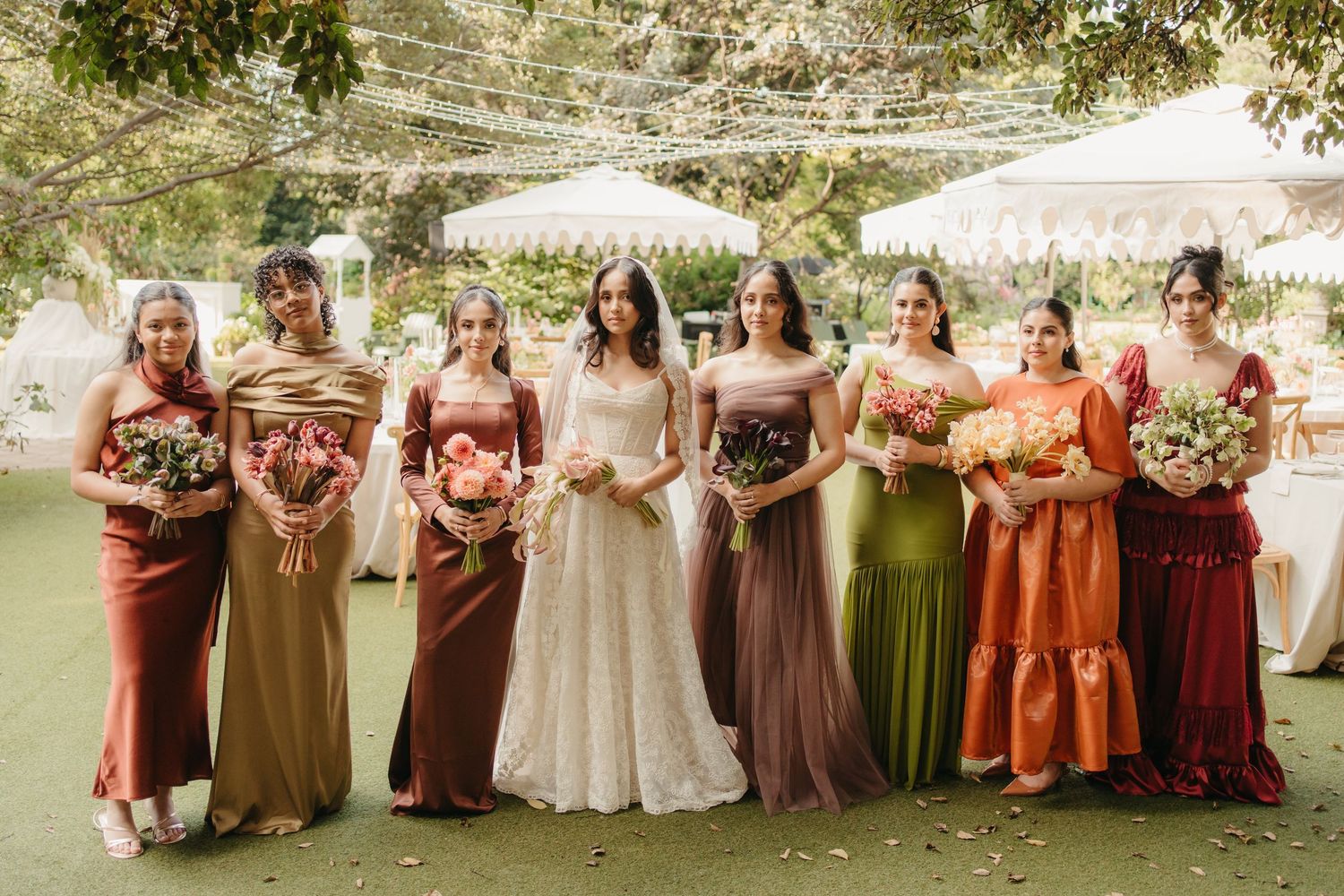 Bride in white lace gown poses with seven bridesmaids in mismatched earth-tone dresses holding bouquets outdoors.