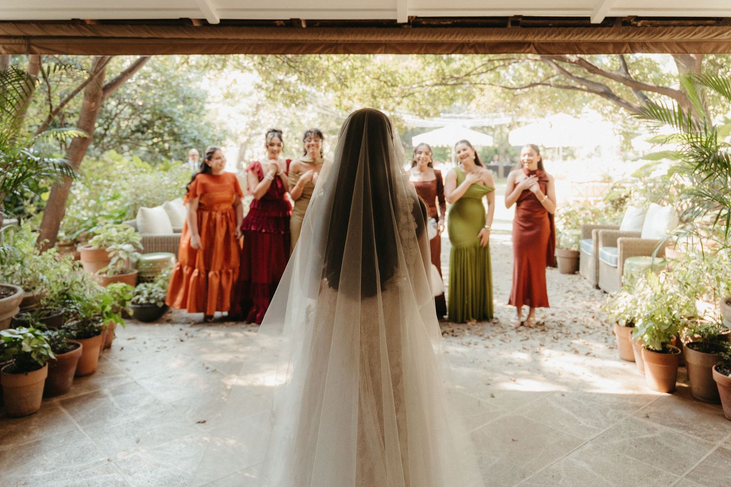 Bride in white gown and veil faces bridesmaids in mixed earth-tone dresses during an outdoor first look.