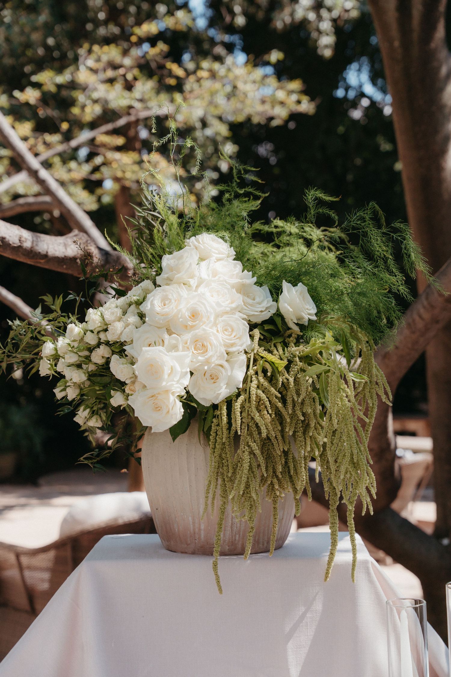 White roses and green ferns in a ceramic vase centerpiece on a white tablecloth at an outdoor wedding.