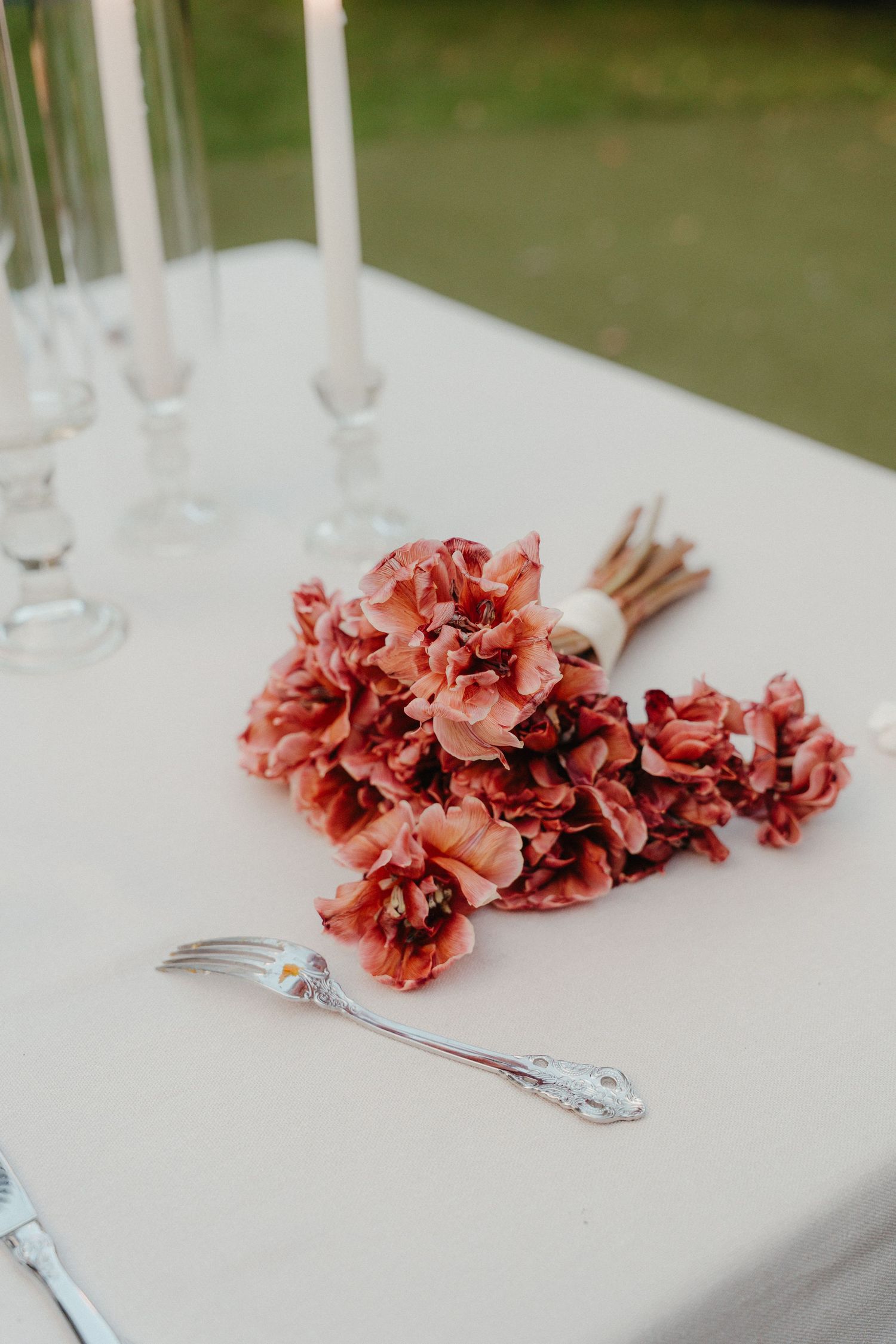 Dusty rose floral bouquet laying on white tablecloth beside silver fork and elegant taper candles at wedding reception.