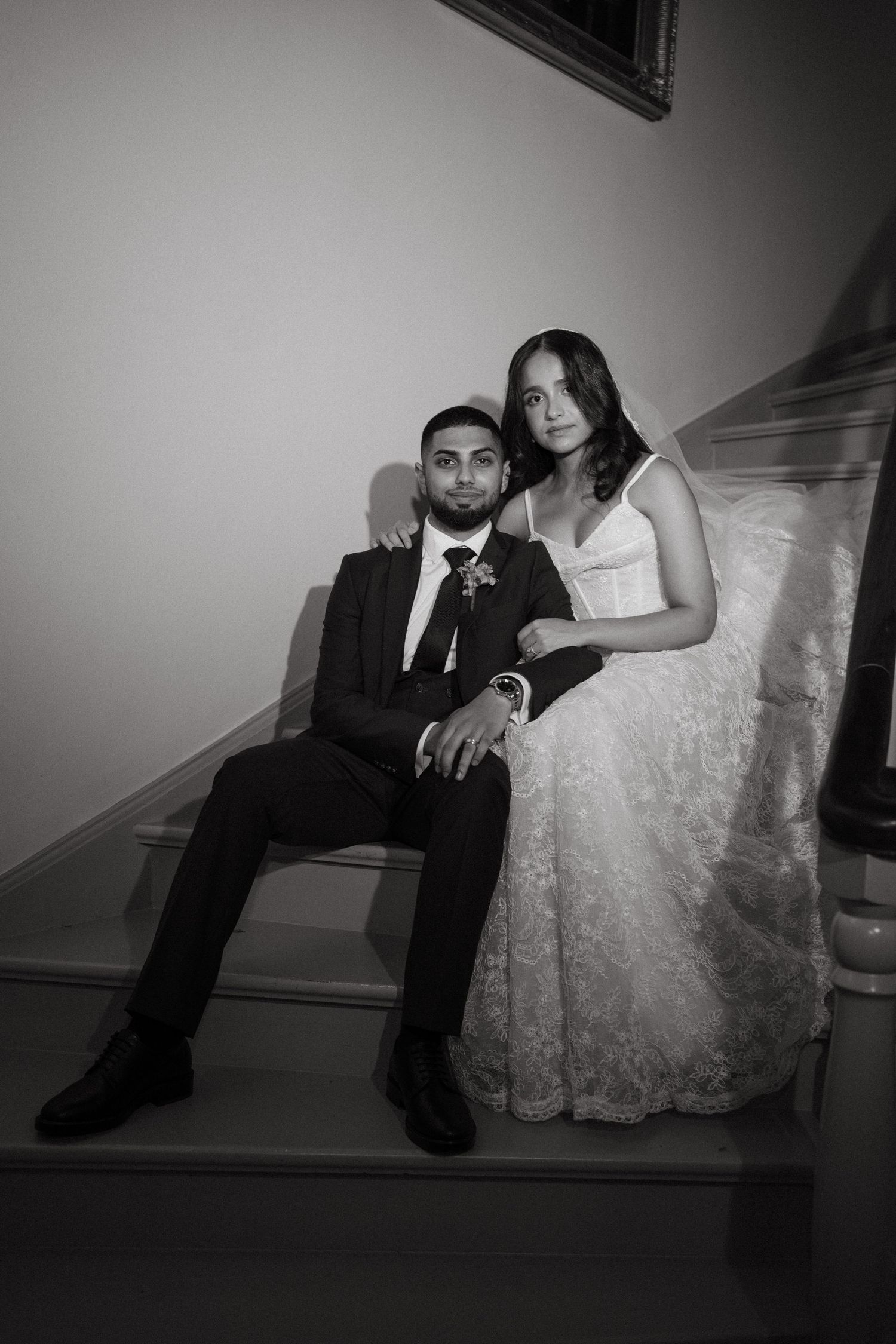 Black and white wedding photo of bride and groom sitting together on staircase, groom in suit, bride in lace gown.