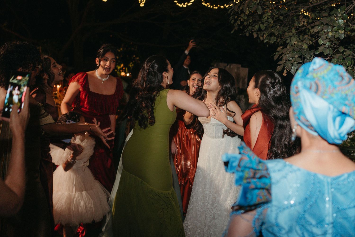 Bride dancing joyfully with guests in colorful dresses at an outdoor evening wedding reception.