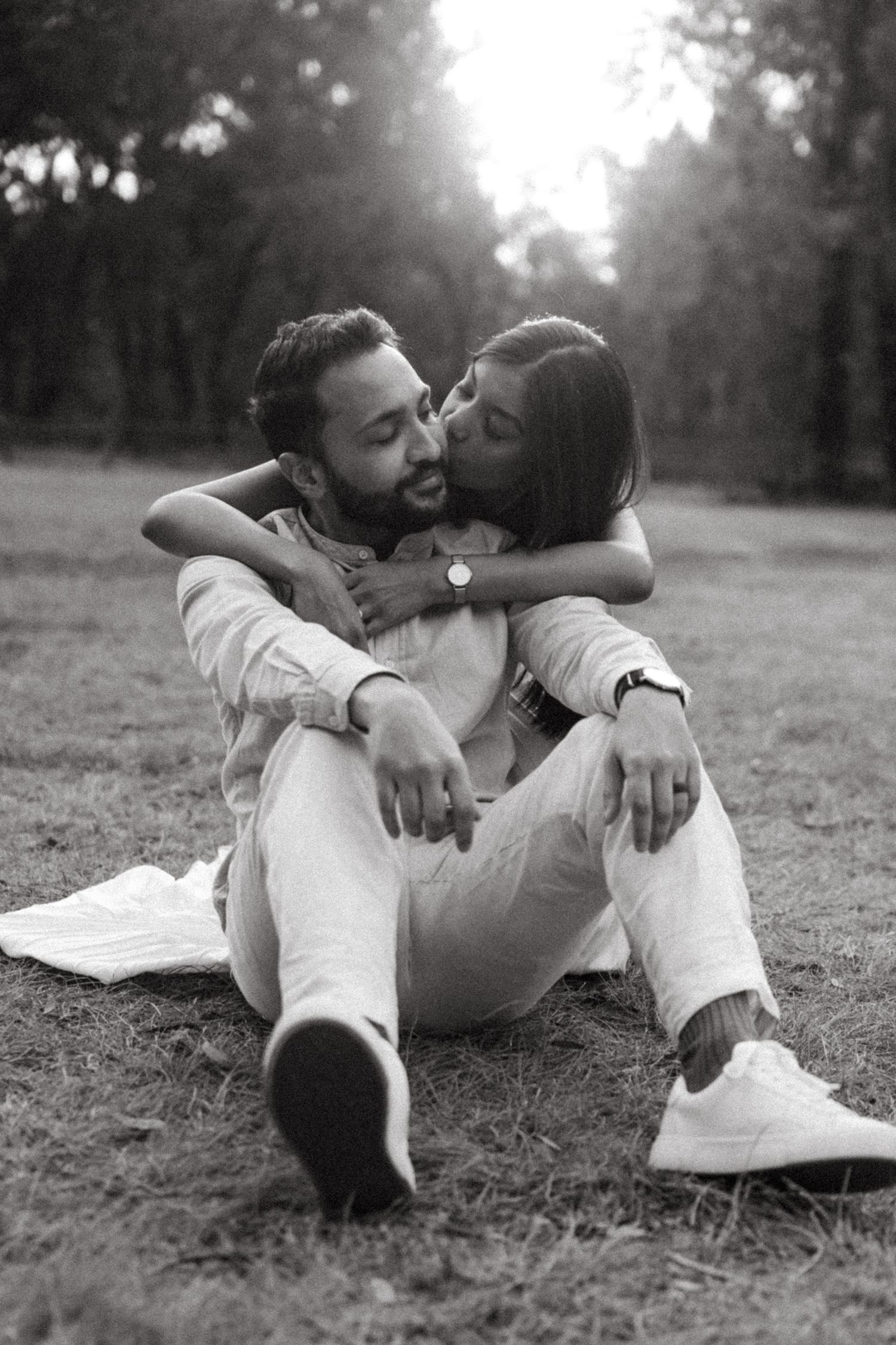 Black and white photo of a woman kissing a seated man on the cheek while hugging him from behind on a grassy park lawn.