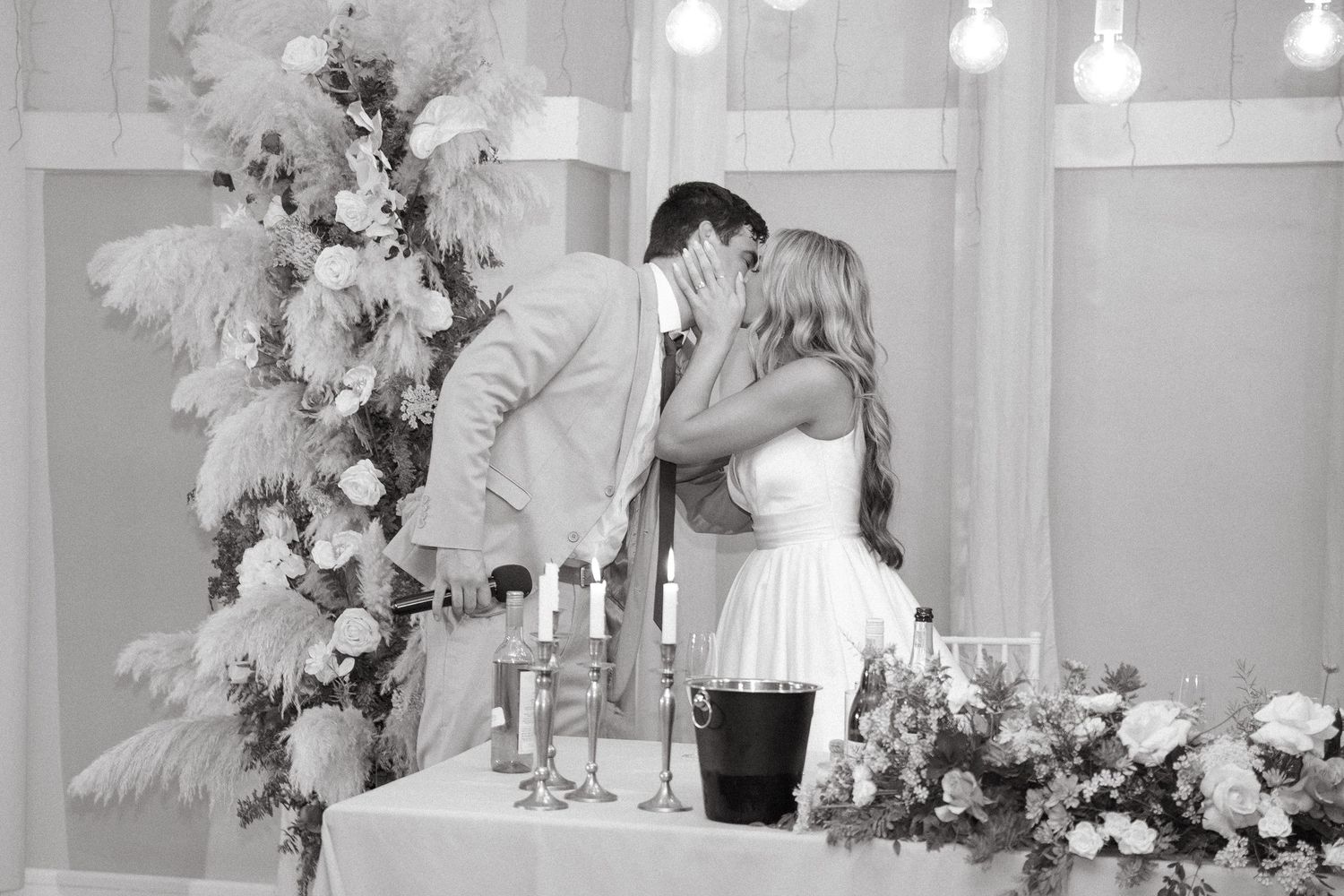 Black and white photo of a bride and groom kissing beside a flower arrangement and a candlelit table with floral centerpieces at a wedding.