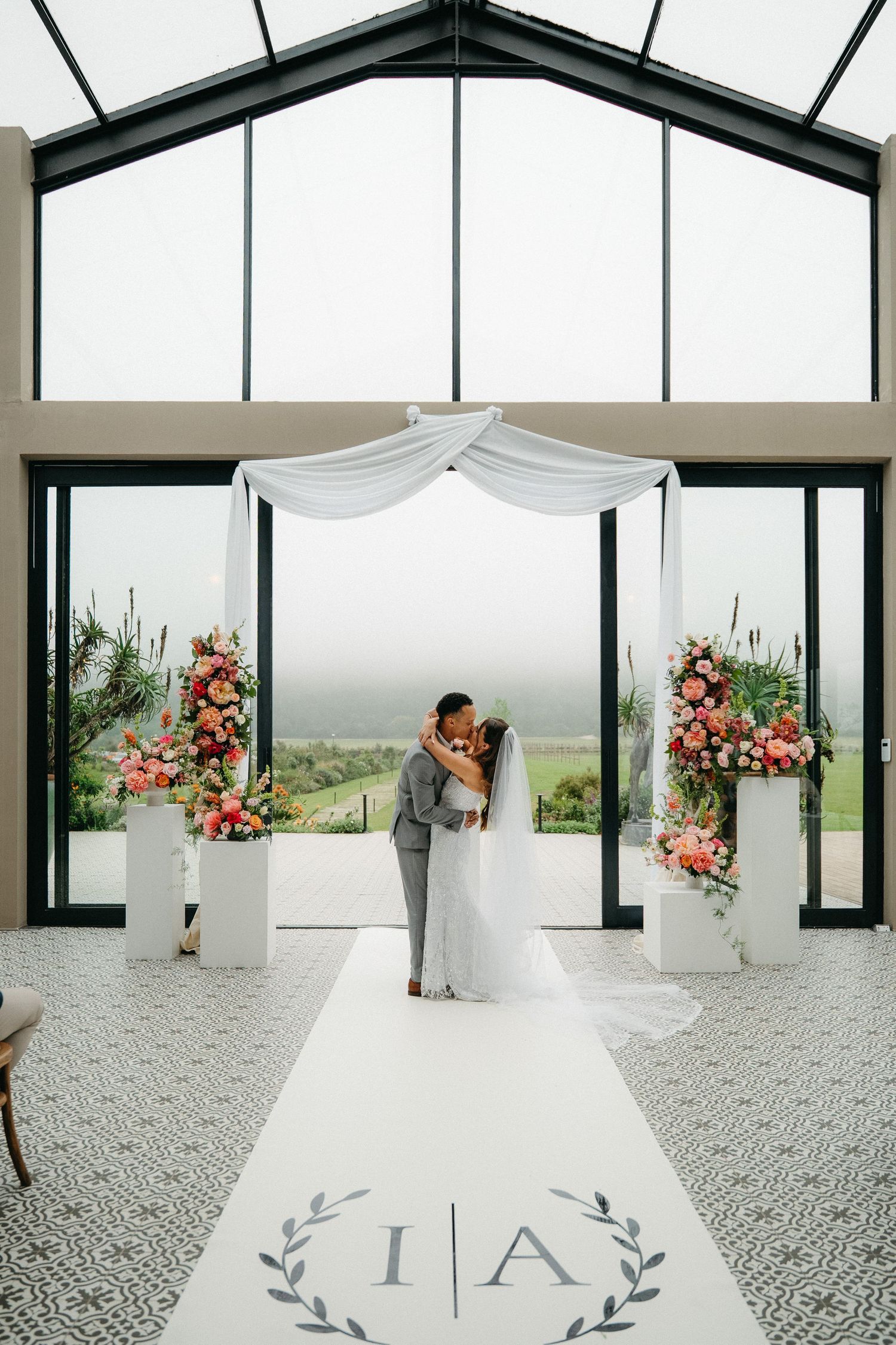 Bride and groom share first kiss at elegant indoor wedding ceremony with floral arrangements and monogrammed aisle runner.