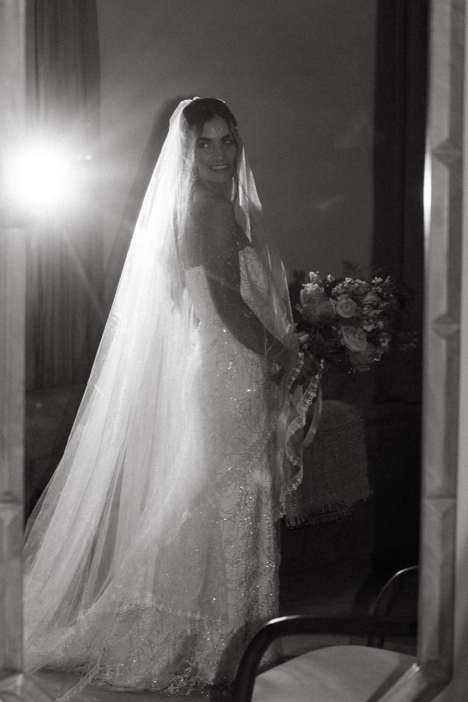 Black and white photo of a bride in a lace gown and long veil, holding a bouquet, smiling over her shoulder.
