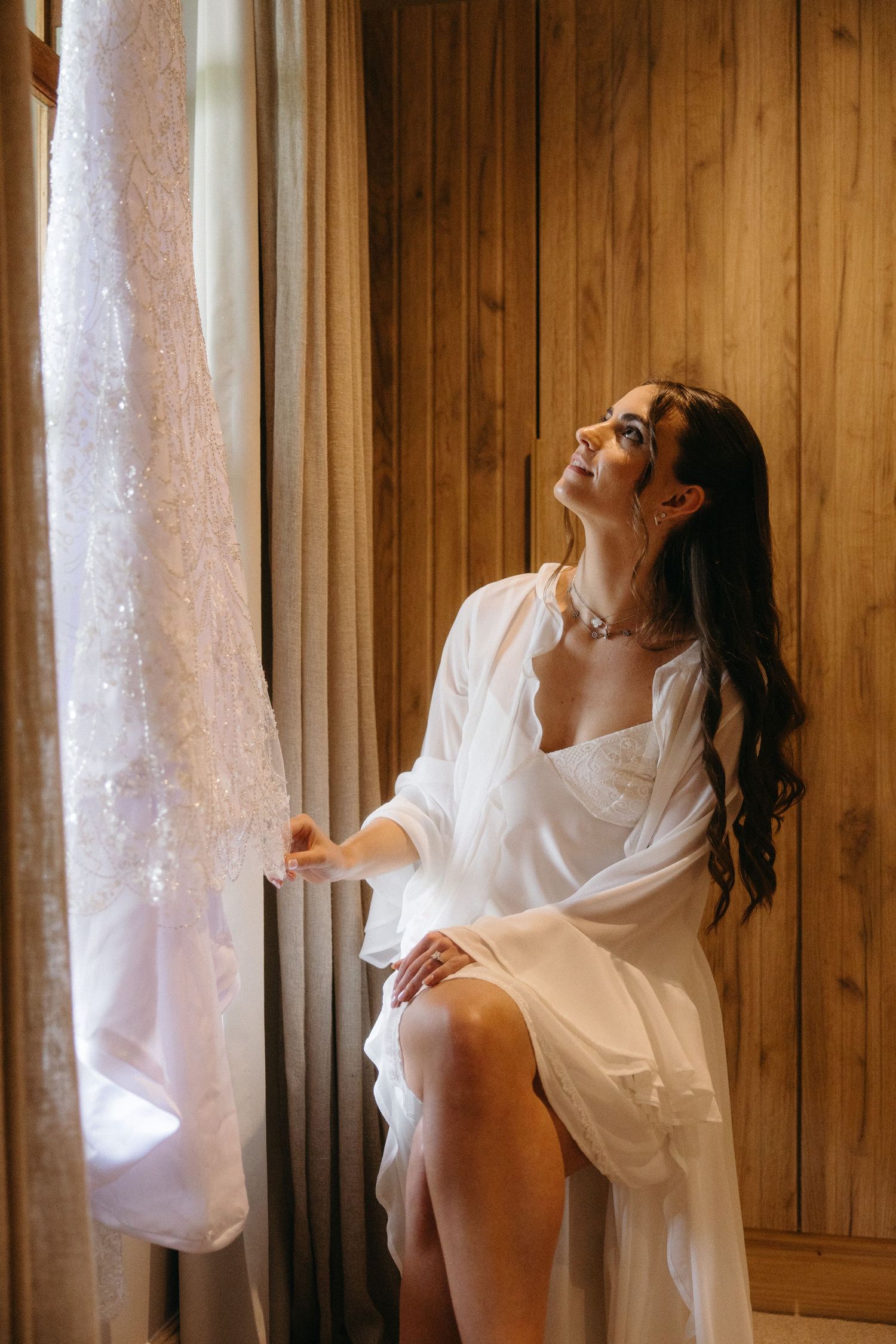 Bride in white satin robe sits by window gazing at her wedding dress in a warm wood-paneled room at Tafika Polo Farm.