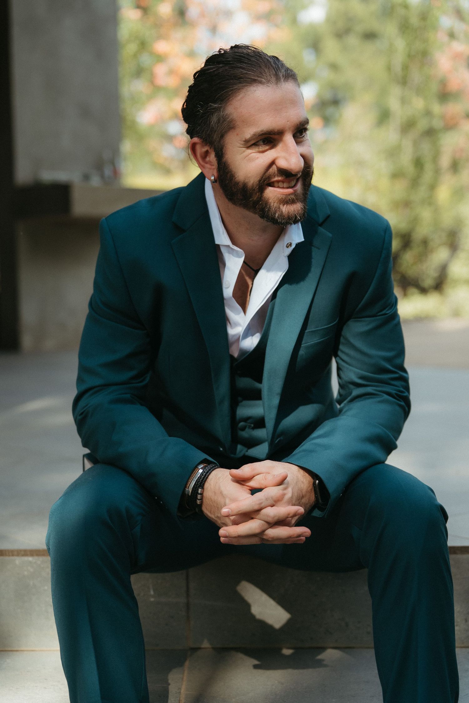 Smiling bearded man in teal three-piece suit sitting outdoors with hands clasped, looking to the side.