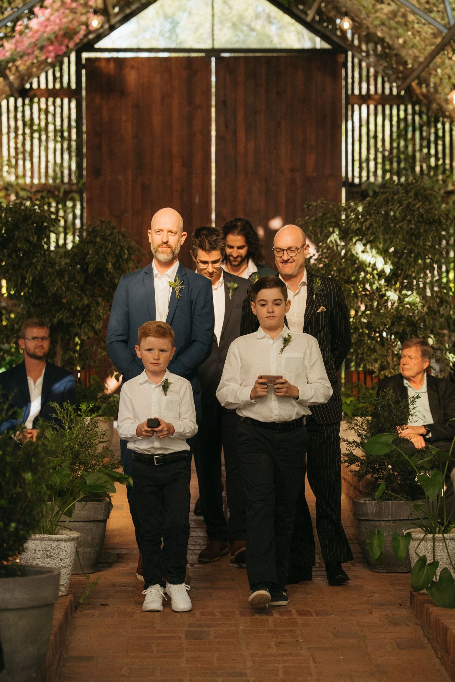 Wedding processional with groomsmen and ring bearers walking down brick aisle through lush greenhouse venue.
