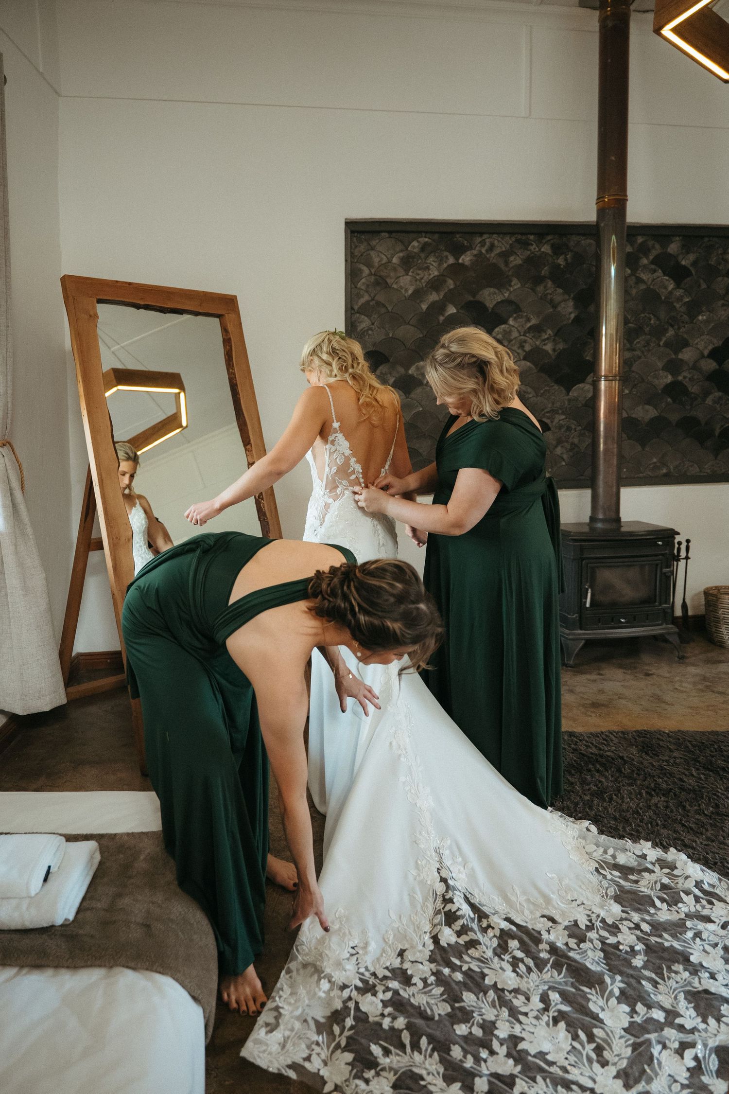 Bridesmaids in green dresses help bride into lace wedding gown before ceremony in rustic room with mirror.