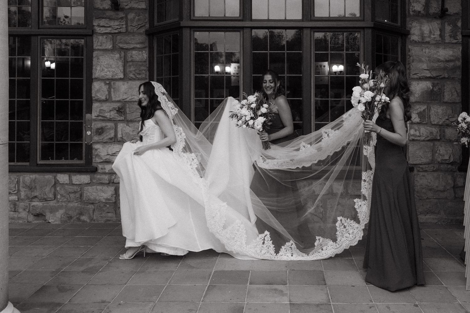Black and white photo of a bride in a white gown with two bridesmaids holding her lace veil outside Glenshiel in Johannesburg