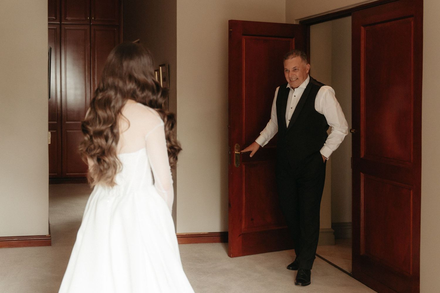 A bride in a white gown shares a first look moment with a man in a black vest and trousers by wooden doors.