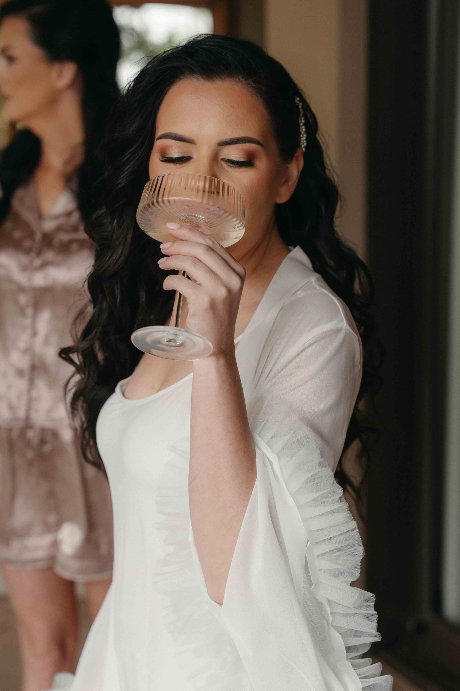 Bride in white dress sips champagne from a coupe glass with eyes closed.