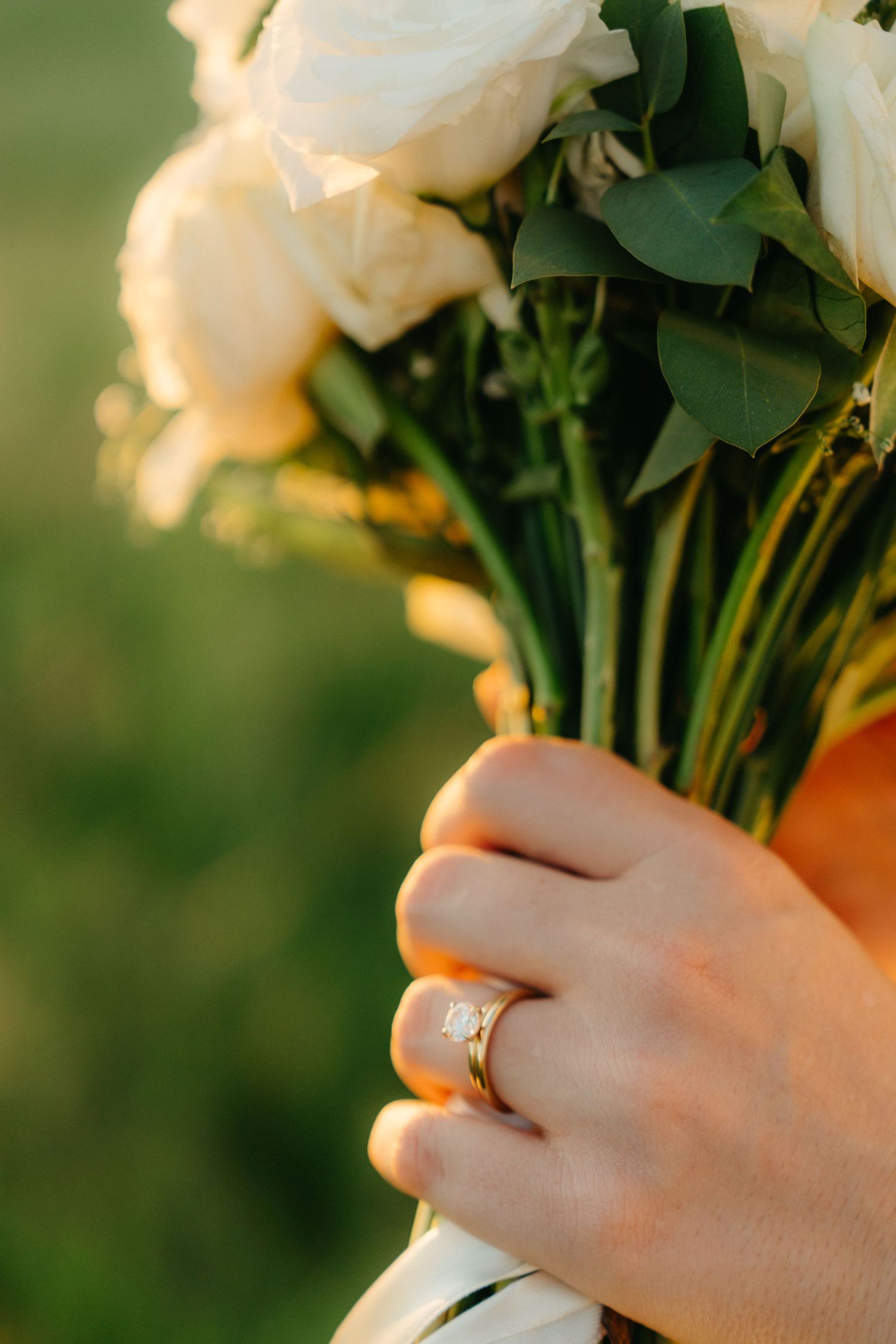 Hand holding white roses in sunlight showing engagement ring and natural bokeh background.