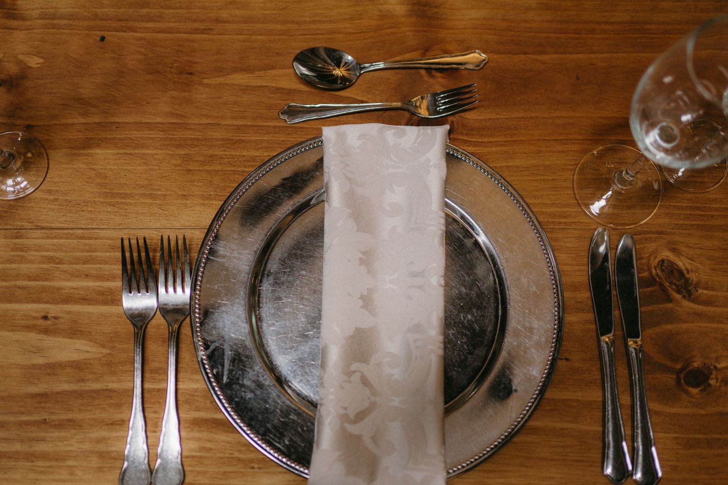 A formal table place setting with an elegant white napkin on a silver plate, surrounded by shining silverware on wood.