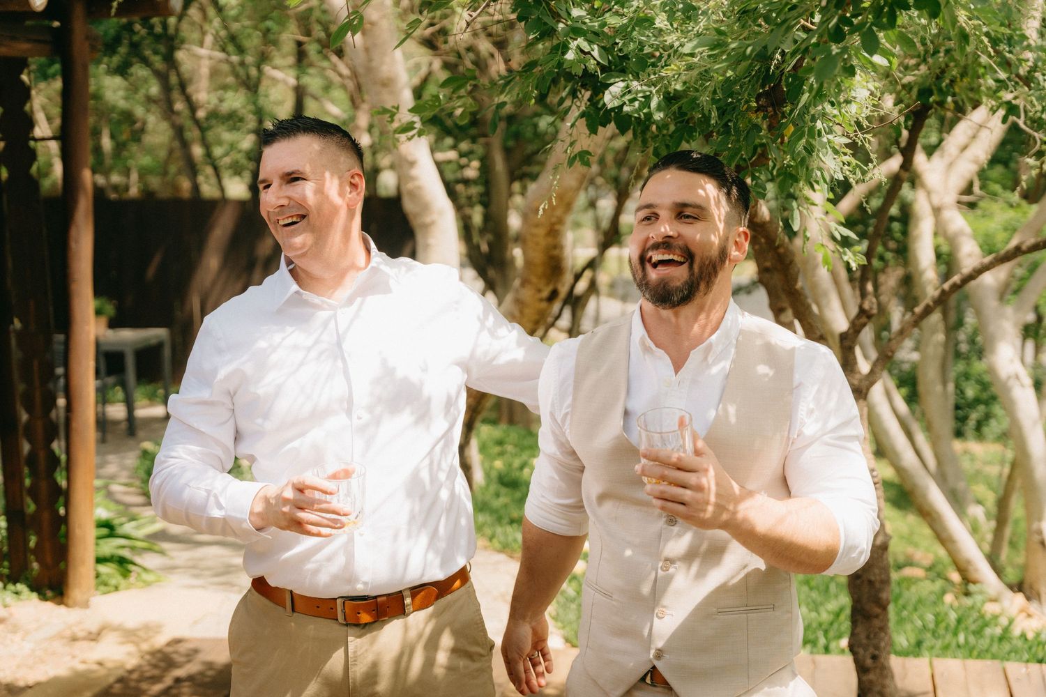 Groomsmen sharing laughs and walking together on a tree-lined path wearing coordinated casual attire.