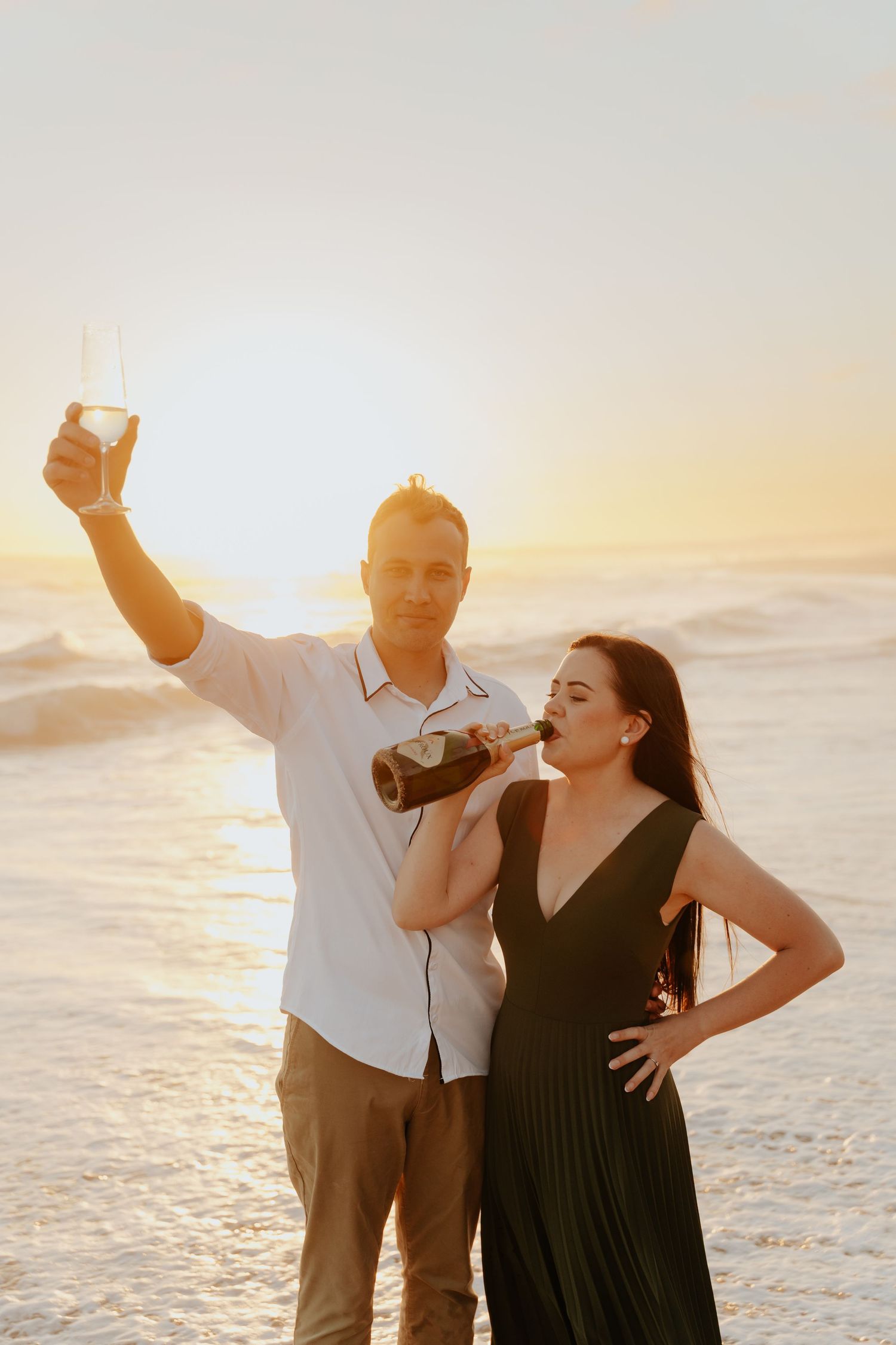 A couple laughs and dances together on the beach at sunset with waves crashing behind them.