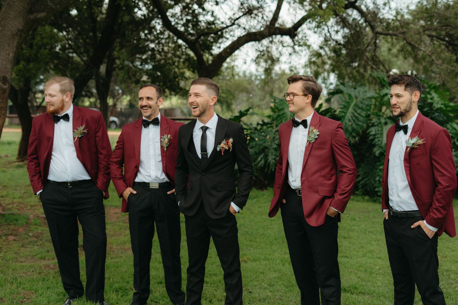 Groomsmen in burgundy suits and black bowties pose confidently outdoors.