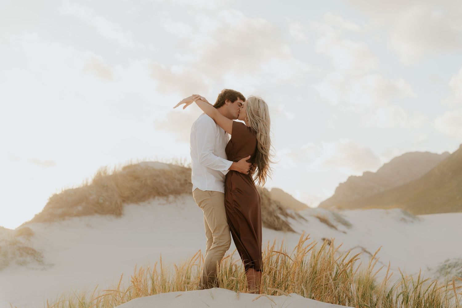 Couple sharing a romantic kiss on sandy dunes with golden grass and soft cloudy sky in a dreamy outdoor setting.