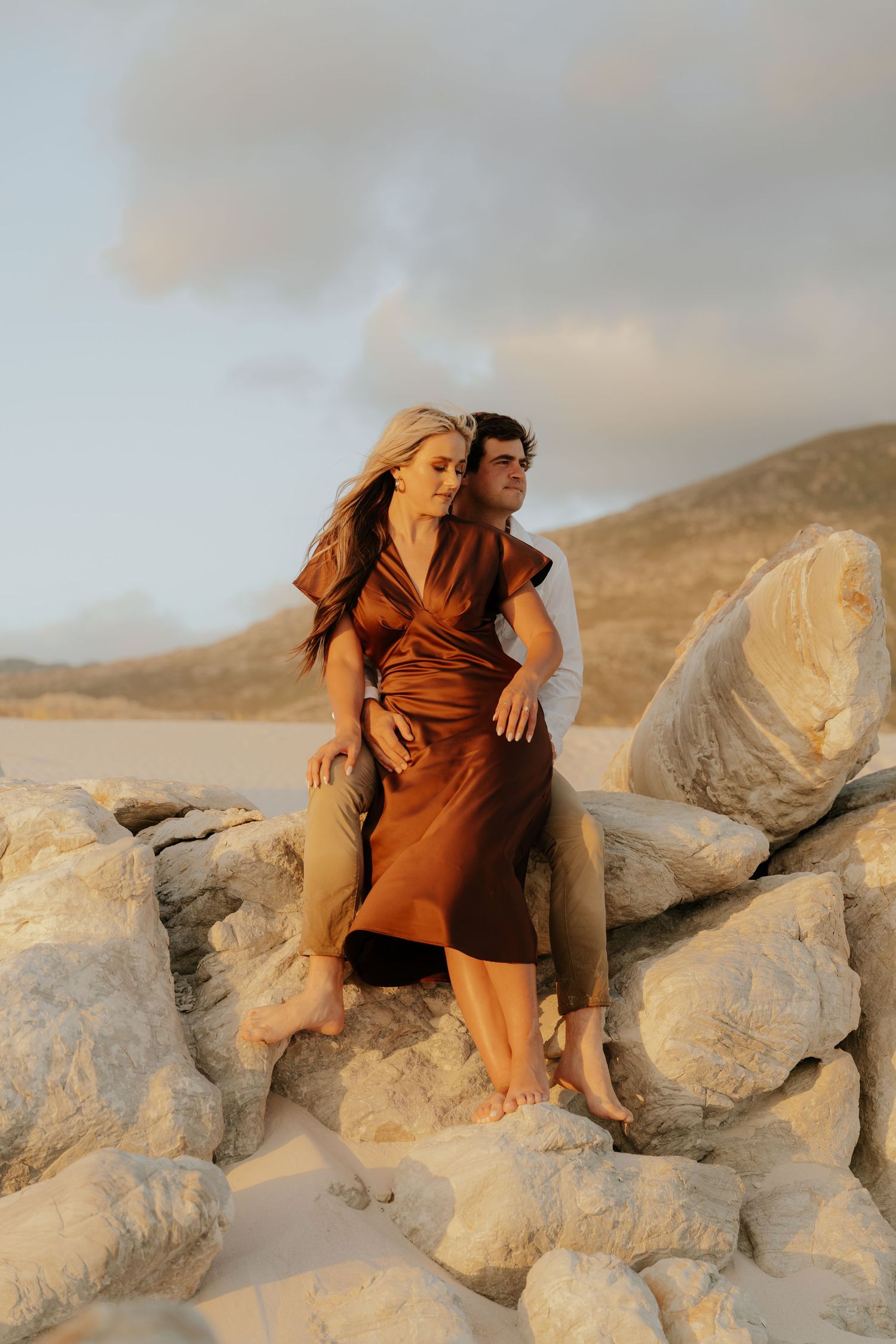Couple sitting on rocks at beach, woman in brown satin dress, golden hour light with sandy dunes and mountains behind.